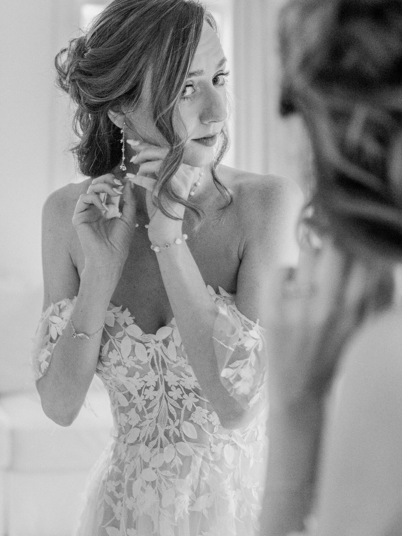 Bride places earrings with a soft smile, reflected in a mirror while wearing her floral lace gown.