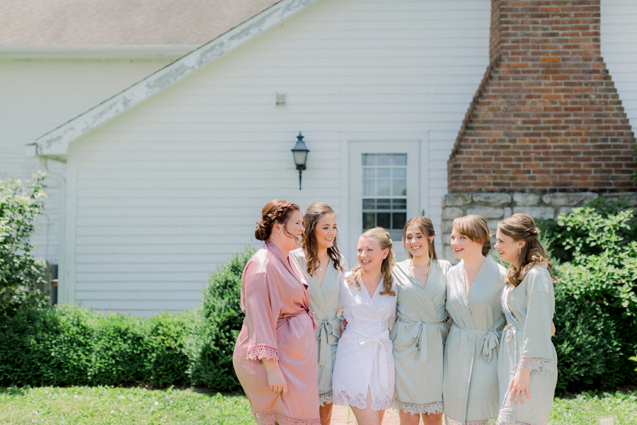 A bride and her bridesmaids laugh together in front of the bridal cottage at a Blue Bell Farm wedding by Love Tree Studios.