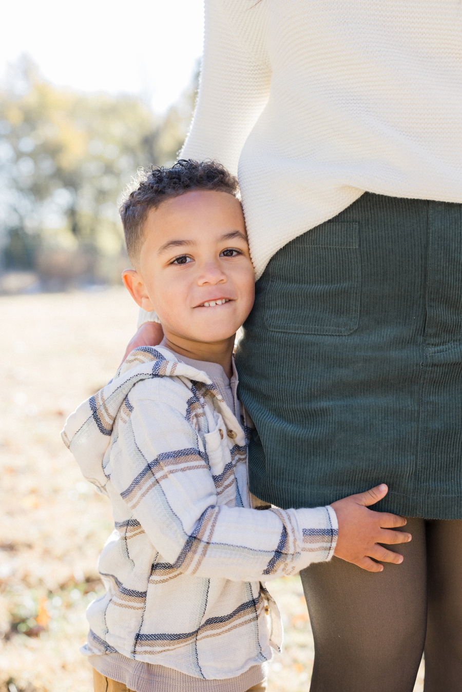 Myles holding onto Mom's leg in the sunlight at Pierpont Farms Family Session, a tender moment captured by Love Tree Studios.