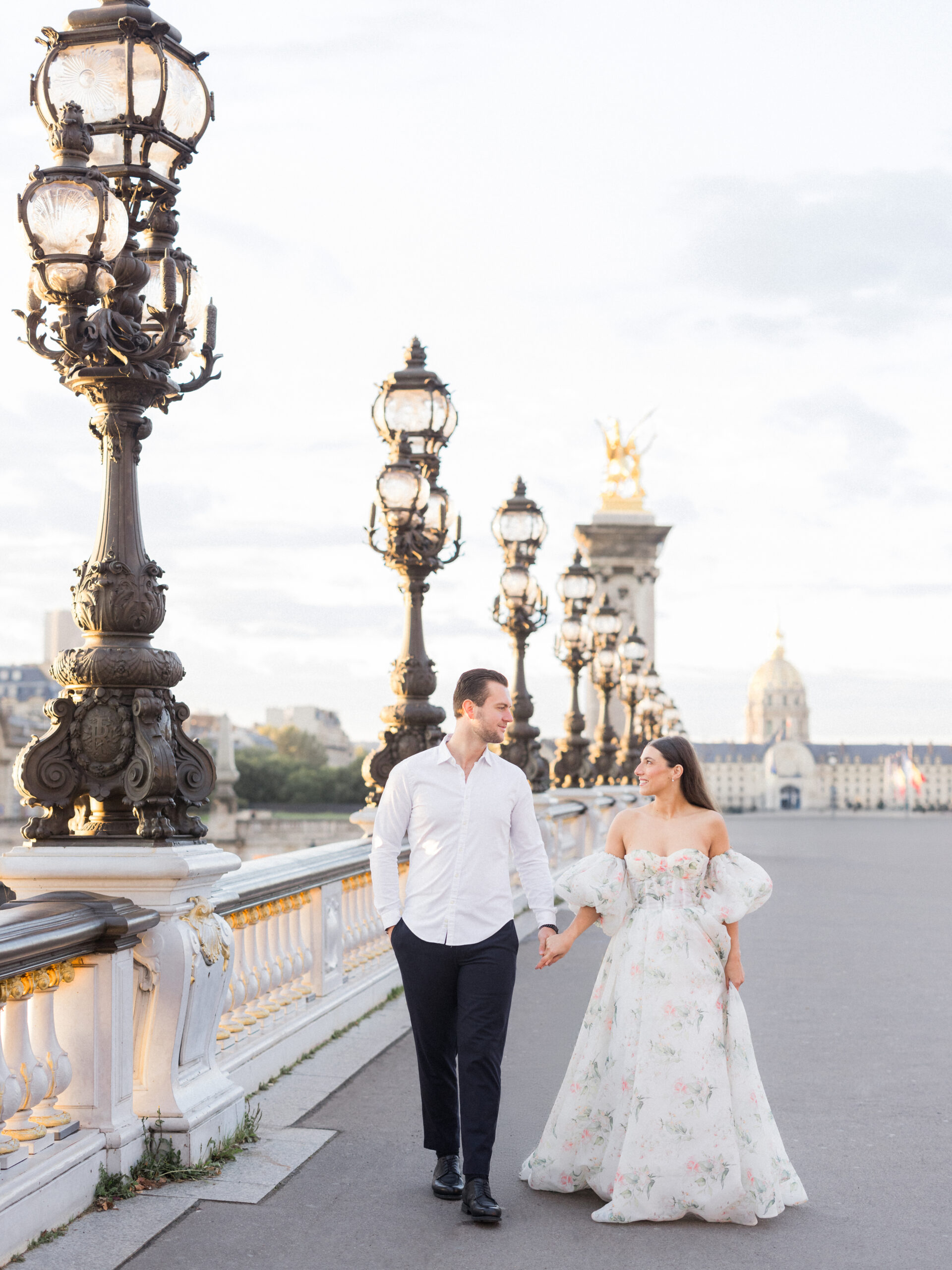 Sinaï and Abraham walking arm in arm along Pont Alexandre III Bridge with Paris landmarks in the distance during their Paris portrait session.