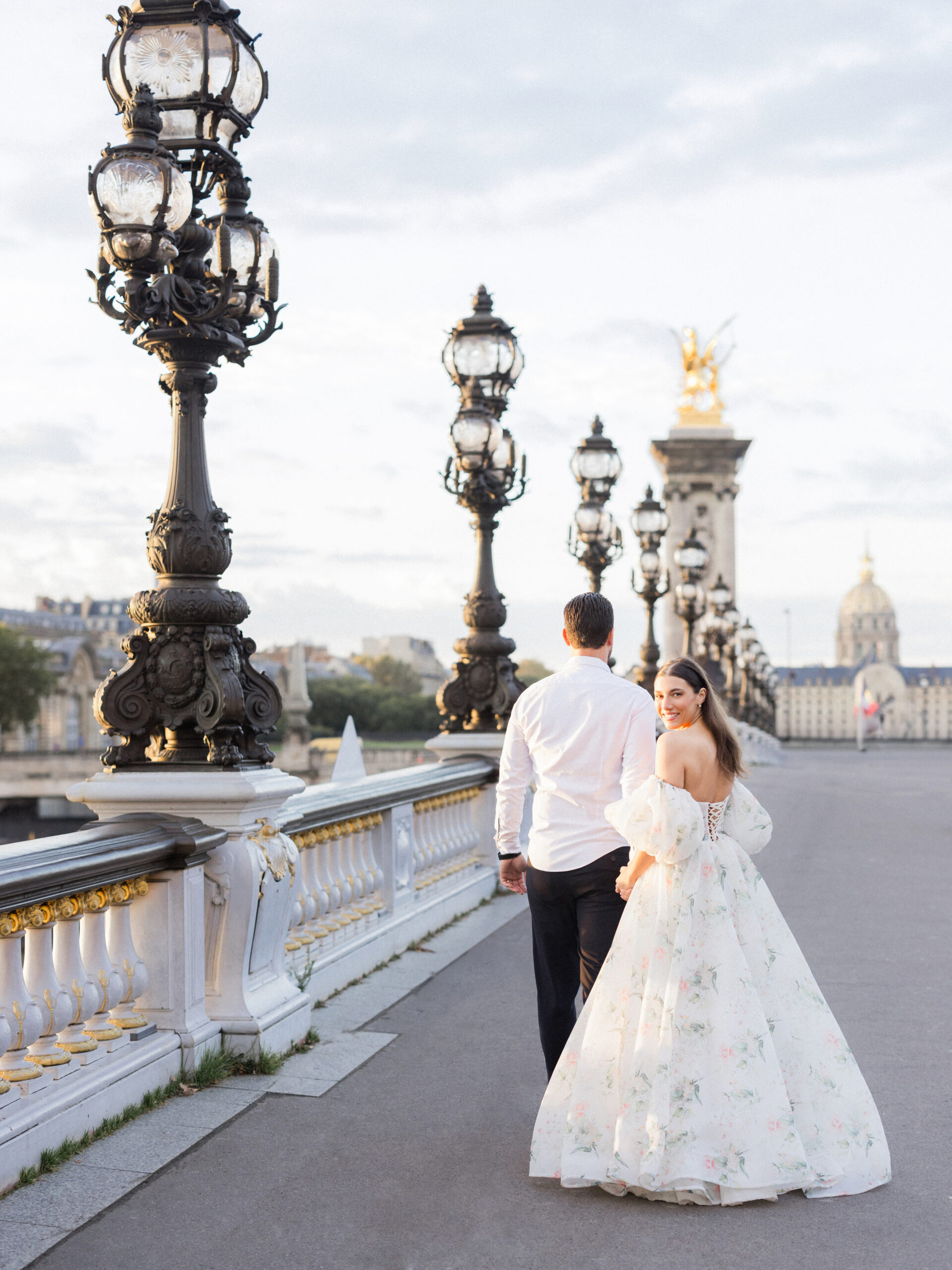 Sinaï and Abraham walking arm in arm along Pont Alexandre III Bridge with Paris landmarks in the distance during their Paris portrait session.