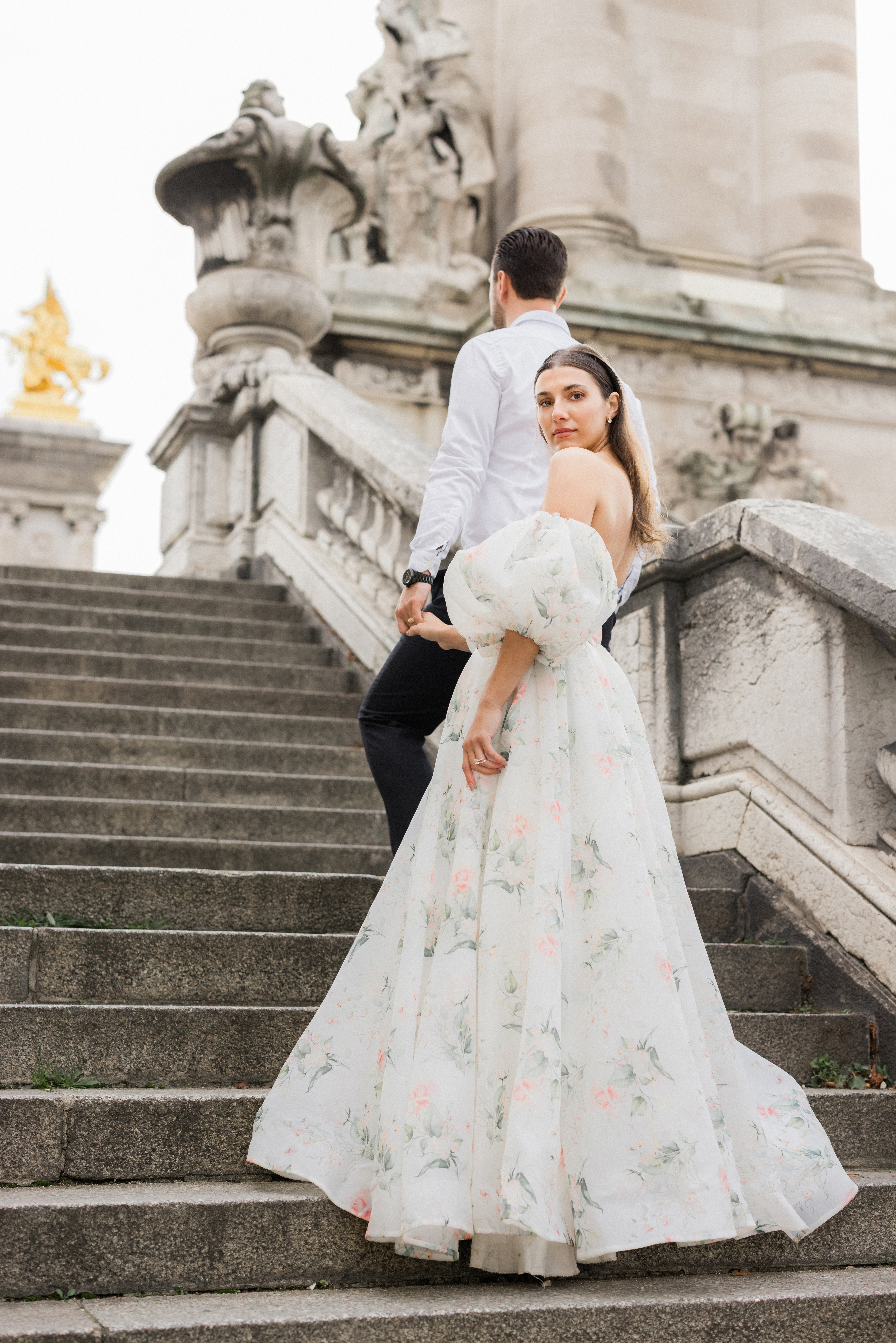 Wide shot of Sinaï and Abraham descending the steps of Pont Alexandre III Bridge, with Paris rooftops in the distance.