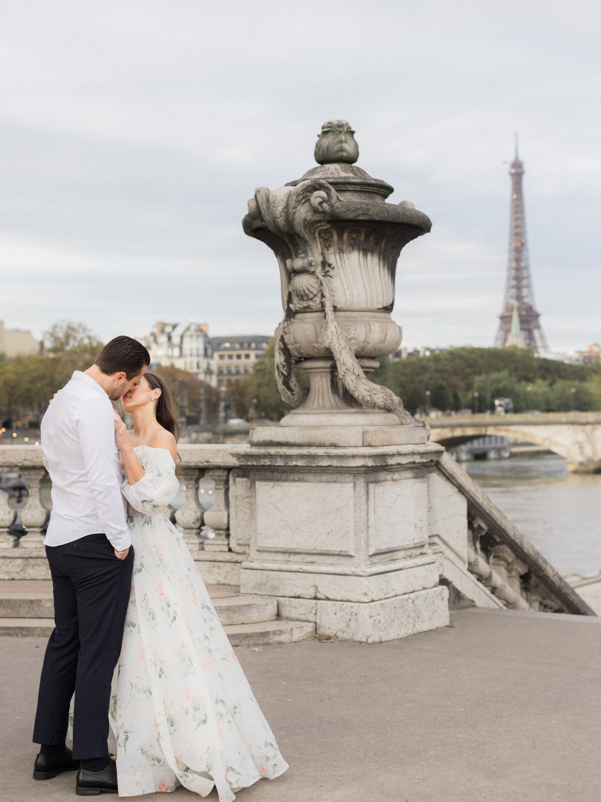 Wide view of Sinaï and Abraham standing near the edge of the bridge, with the Eiffel Tower visible in the distance during their Paris portrait session Love Tree Studios.