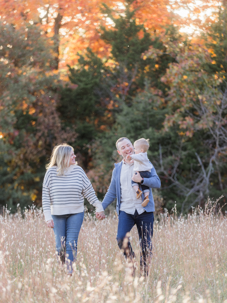 Father holding baby while mother smiles outdoors in vibrant fall scenery, photographed by Love Tree Studios in Columbia, MO.