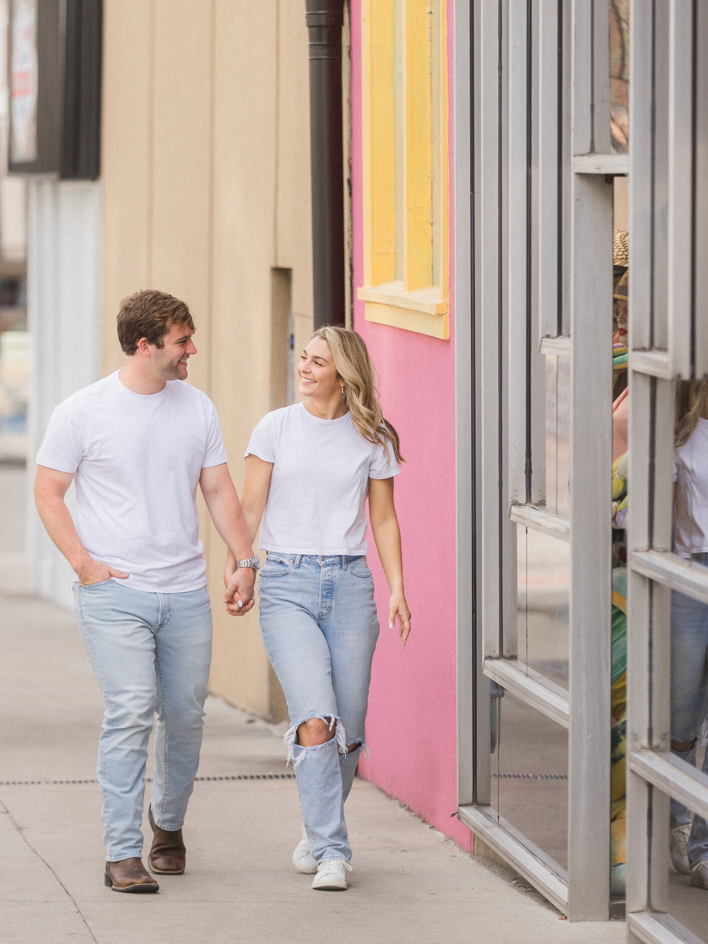 Engaged couple walking hand-in-hand down a colorful downtown sidewalk in casual outfits.