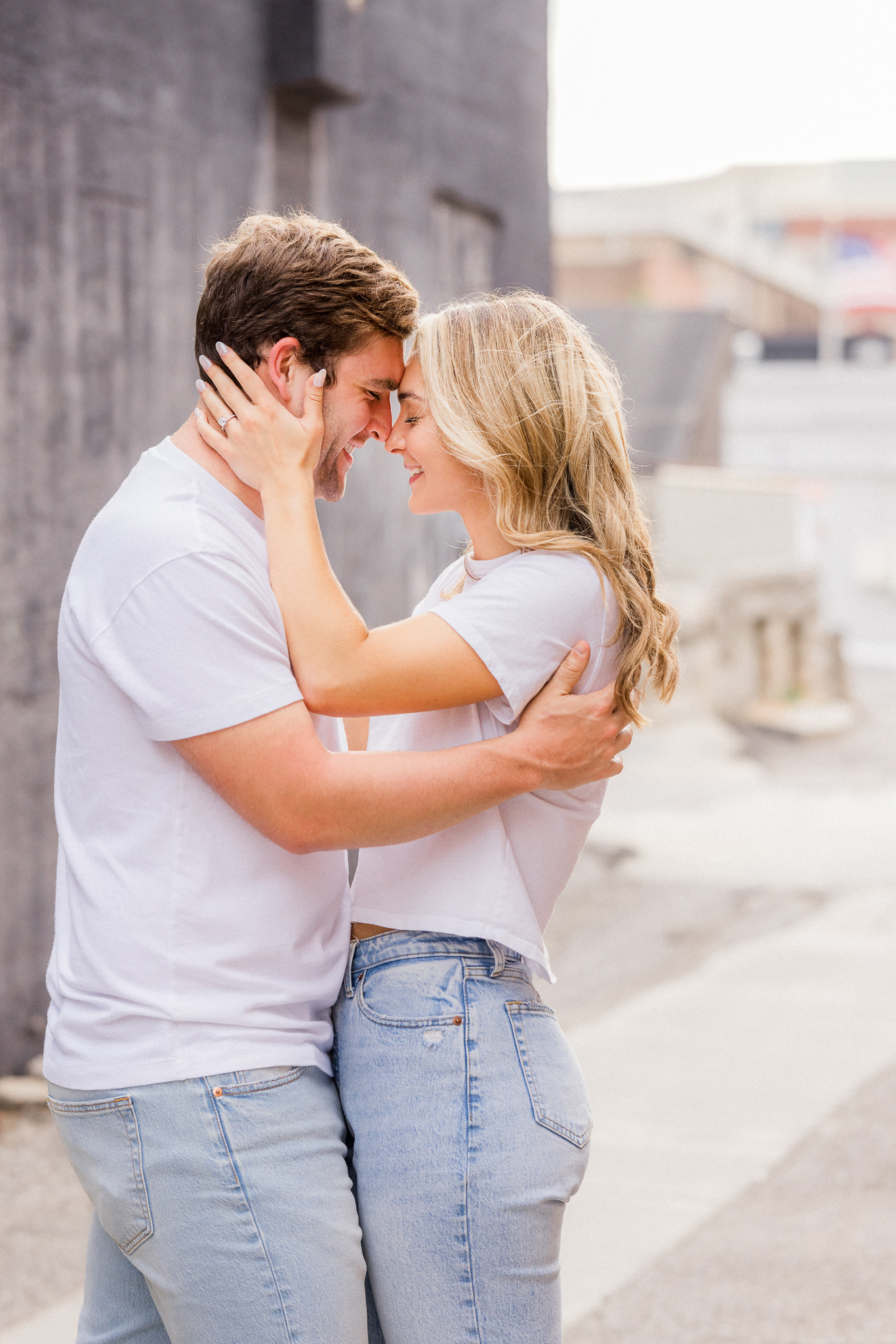 Groom-to-be laughing as bride-to-be gently cups his face during their downtown spring engagement session.