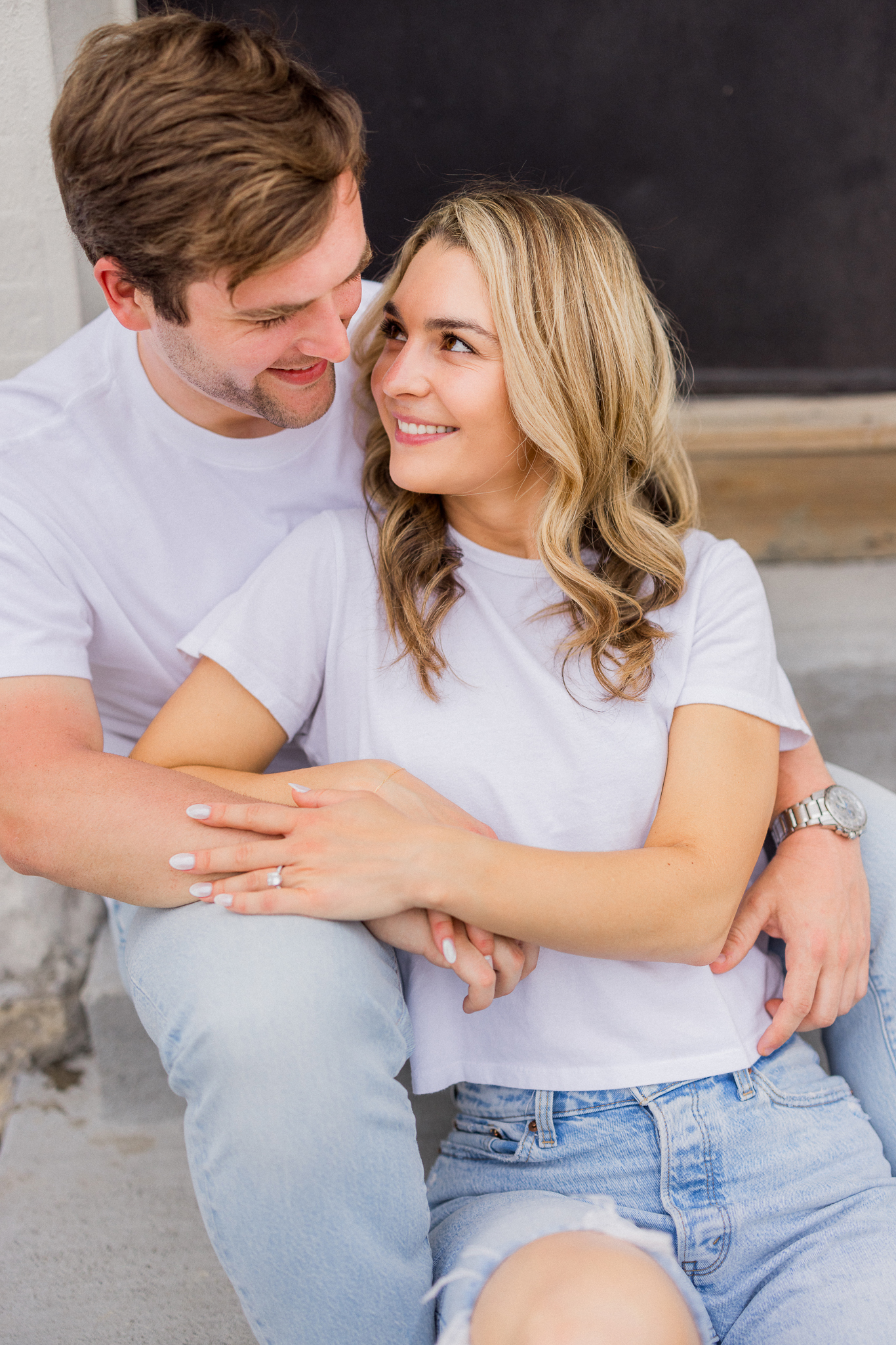 Close-up of engaged couple sitting on concrete steps, smiling and snuggled close in matching white shirts.