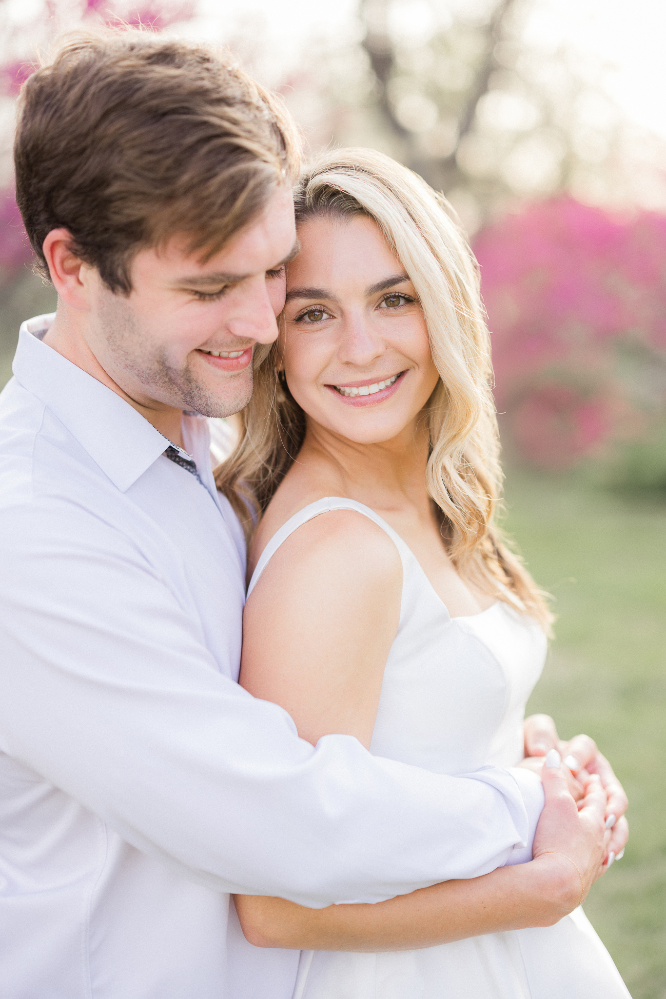 Bride-to-be smiling at the camera while groom-to-be hugs her from behind under vibrant pink blossoms.