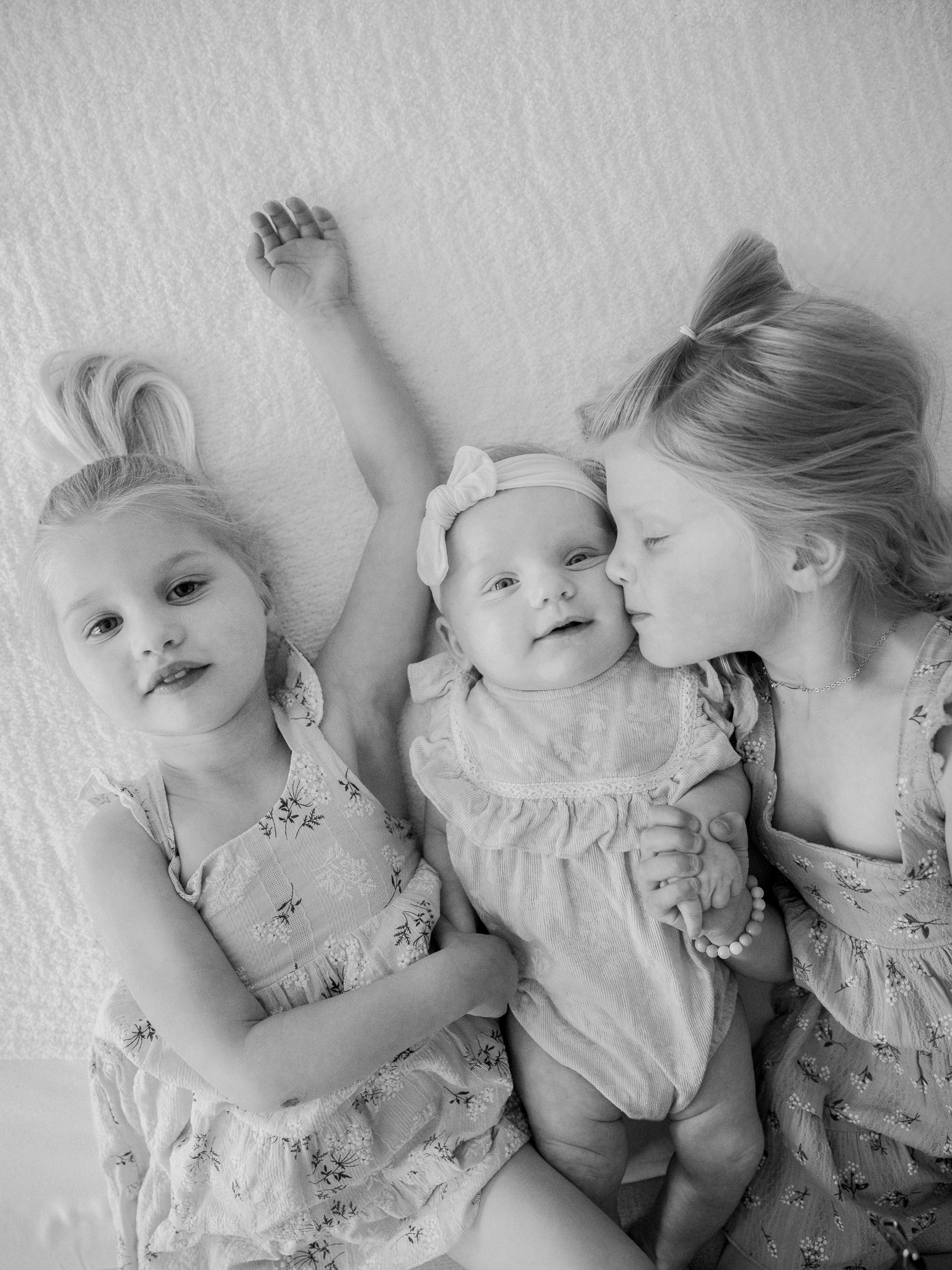 Black and white photo of three sisters lying on a bed during an in-home newborn photography session in Missouri with Love Tree Studios.