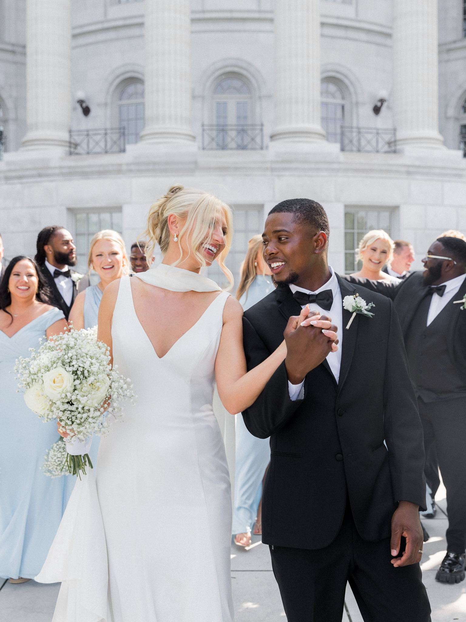 Bride and groom hold hands and smile at each other, walking together with the bridal party behind them.