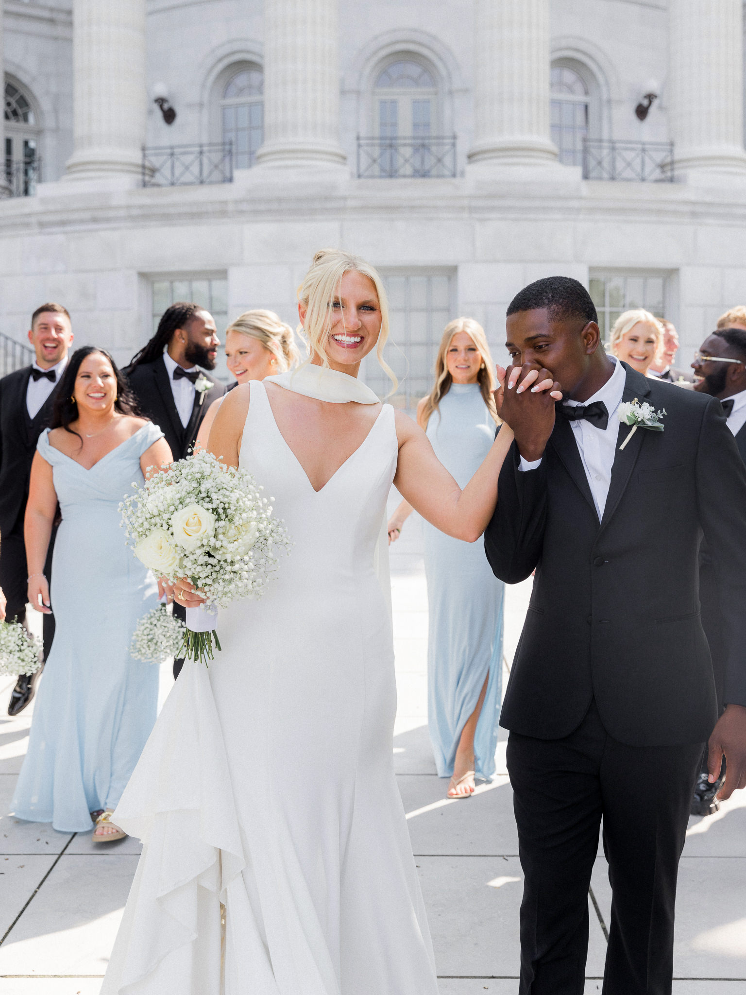 Romantic moment of the groom kissing the bride’s hand as she beams, surrounded by their wedding party.