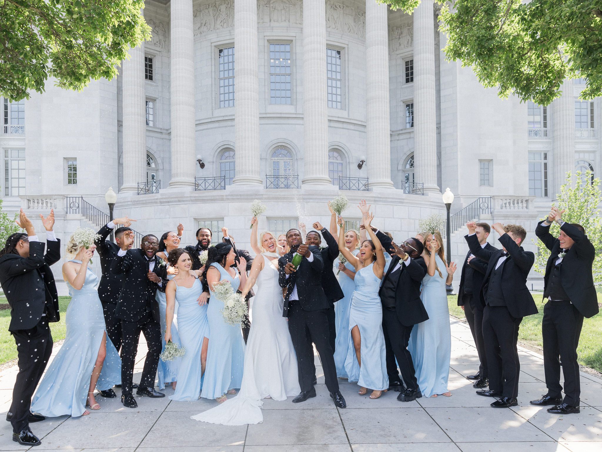 Large wedding party pop champagne outside a grand white style building.
