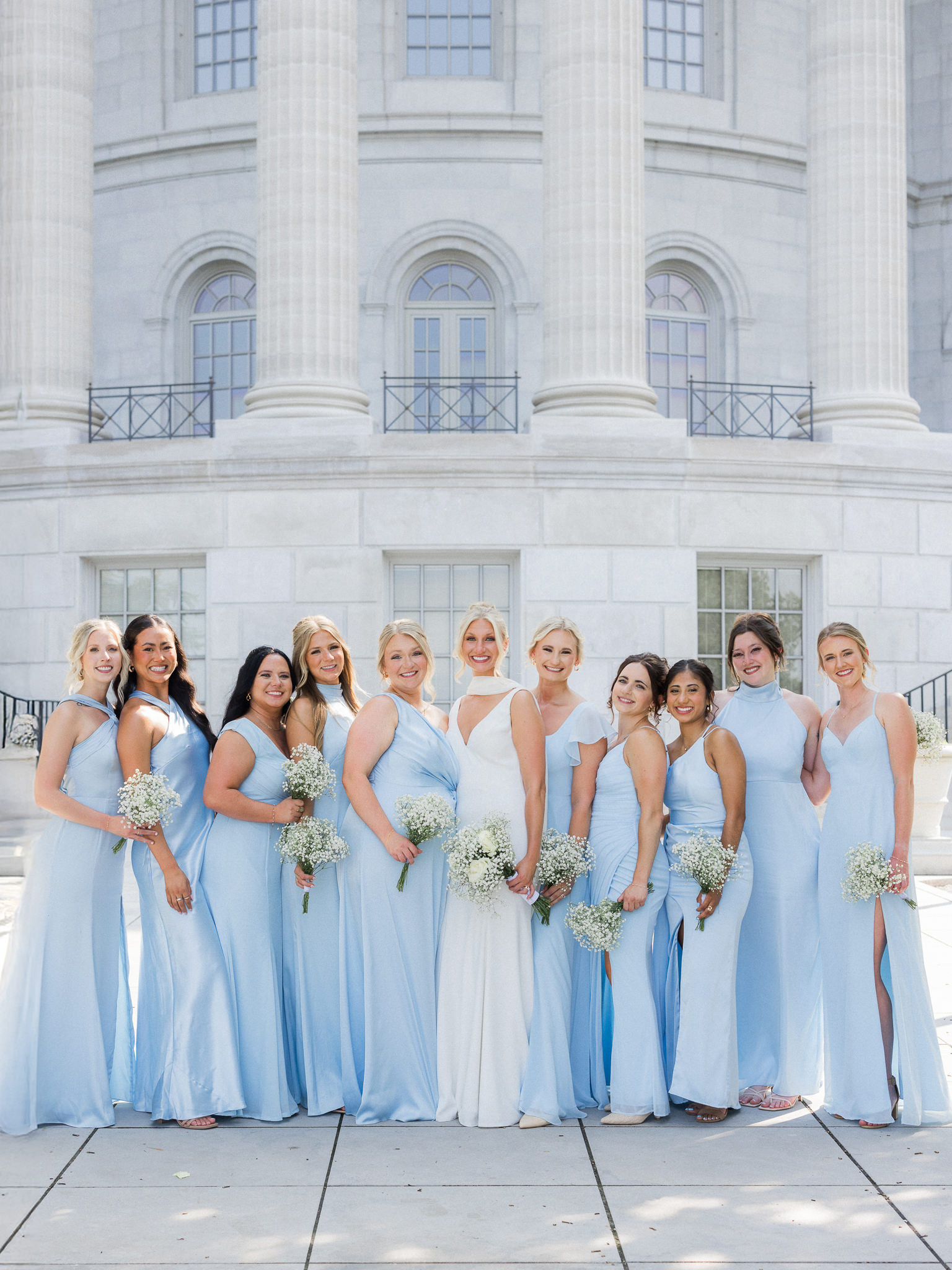 ride surrounded by bridesmaids in light blue dresses, all holding baby's breath bouquets, in front of a neoclassical building.