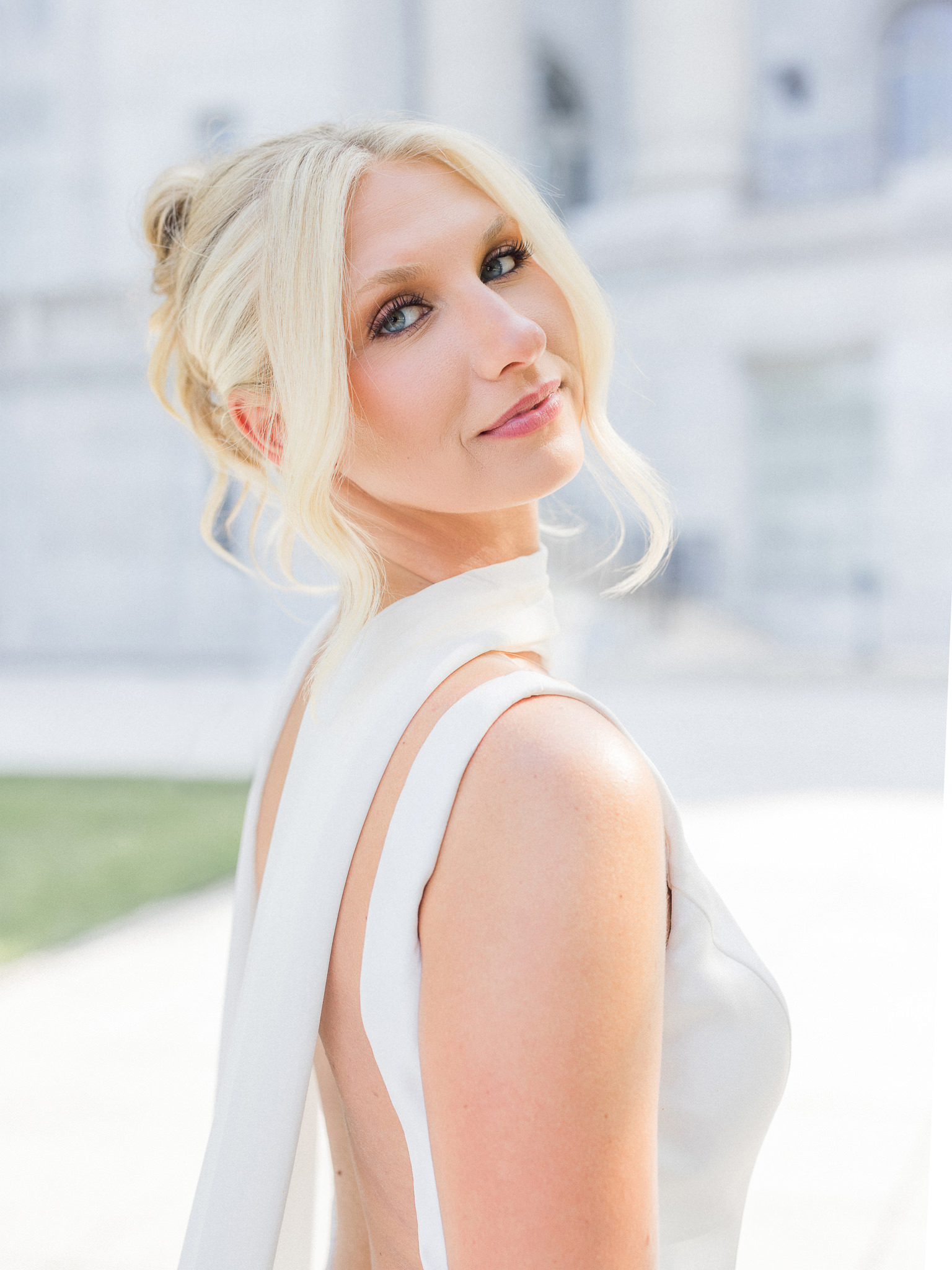 Blonde bride with soft curls smiles over her shoulder in a sleek white gown during a sunny outdoor bridal portrait.