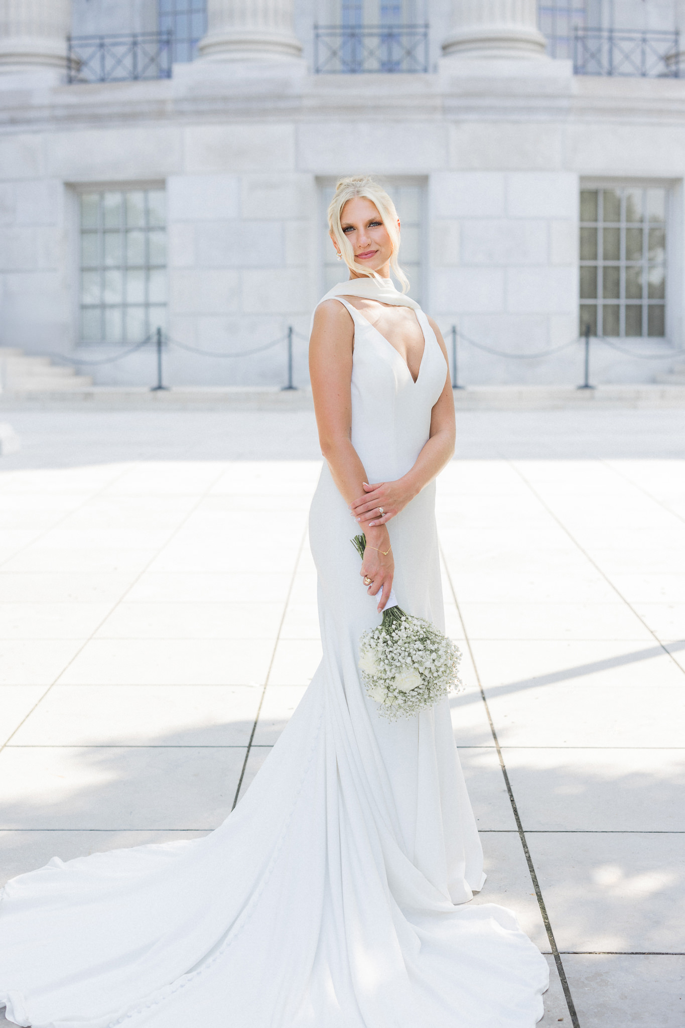 Elegant bride in a minimalist white gown holds a bouquet of baby's breath and white roses, standing on a grand marble terrace.