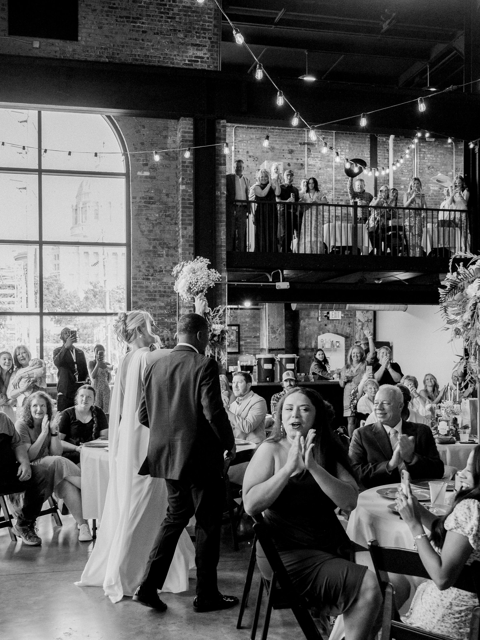Bride and groom make a grand entrance into their reception space, with guests clapping and cheering in a warmly lit industrial venue.