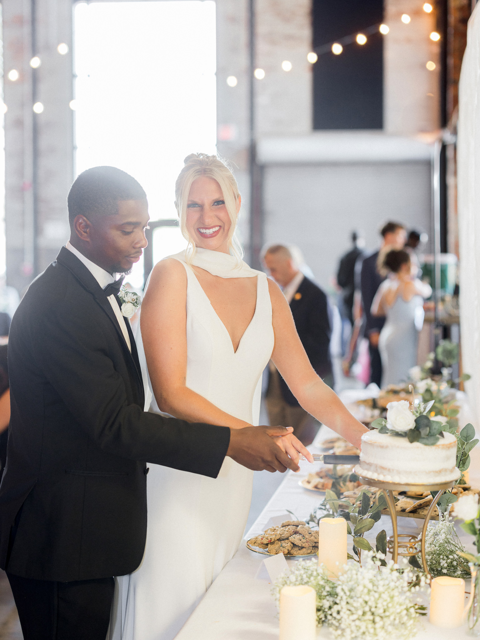 Bride and groom smile while cutting their wedding cake, surrounded by desserts and glowing candlelight at the reception.