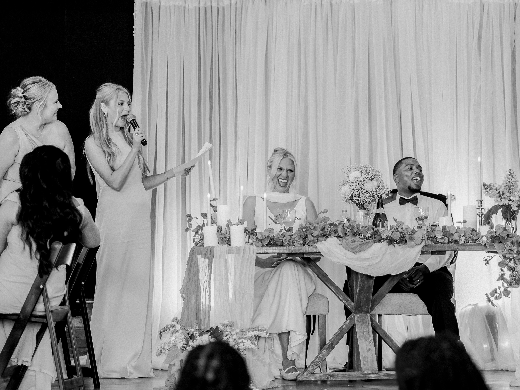 Bridesmaid delivers a speech while the bride and groom laugh during the reception.