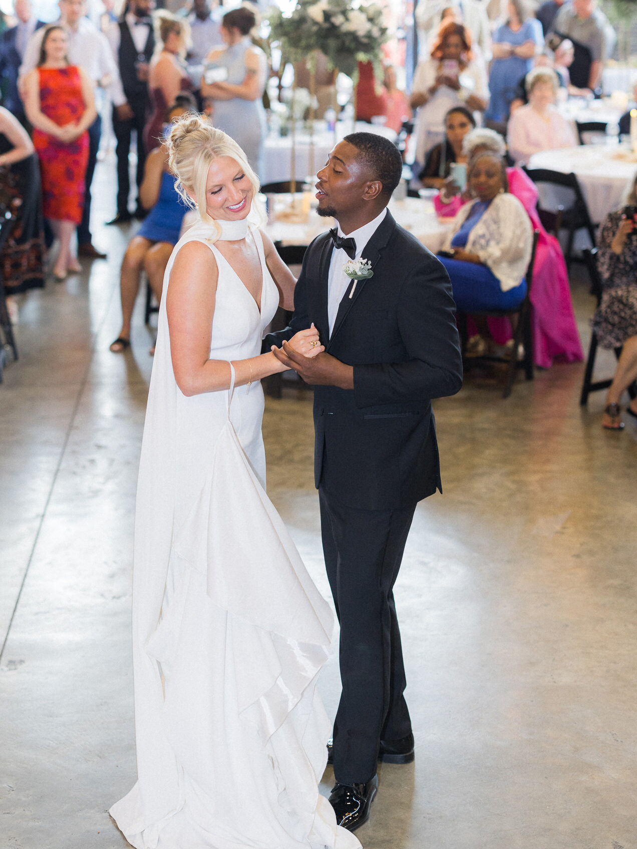 Groom gazes at bride as they enjoy their first dance with wedding guests watching.