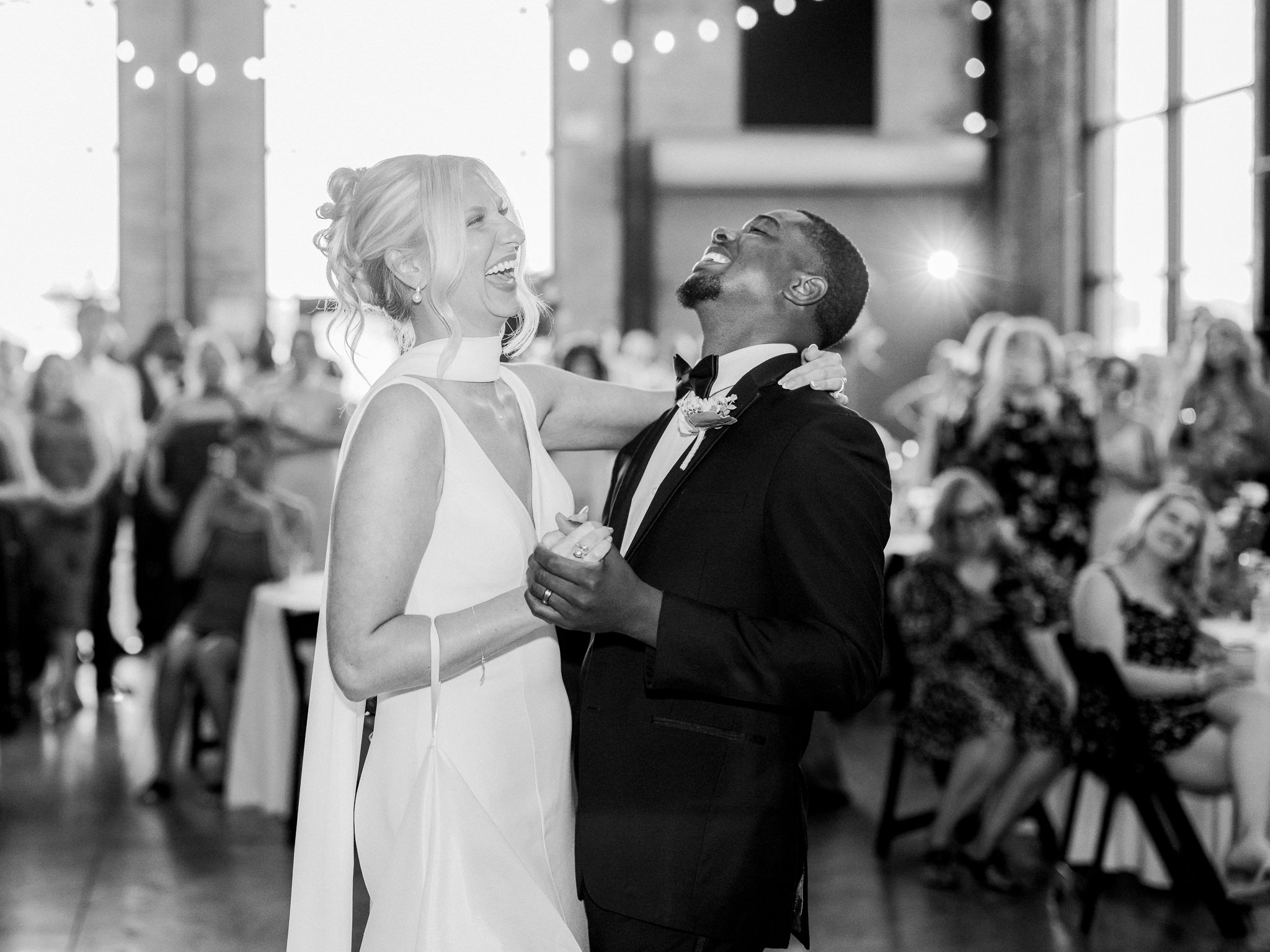 Bride and groom laugh together during their first dance, surrounded by joyful guests.