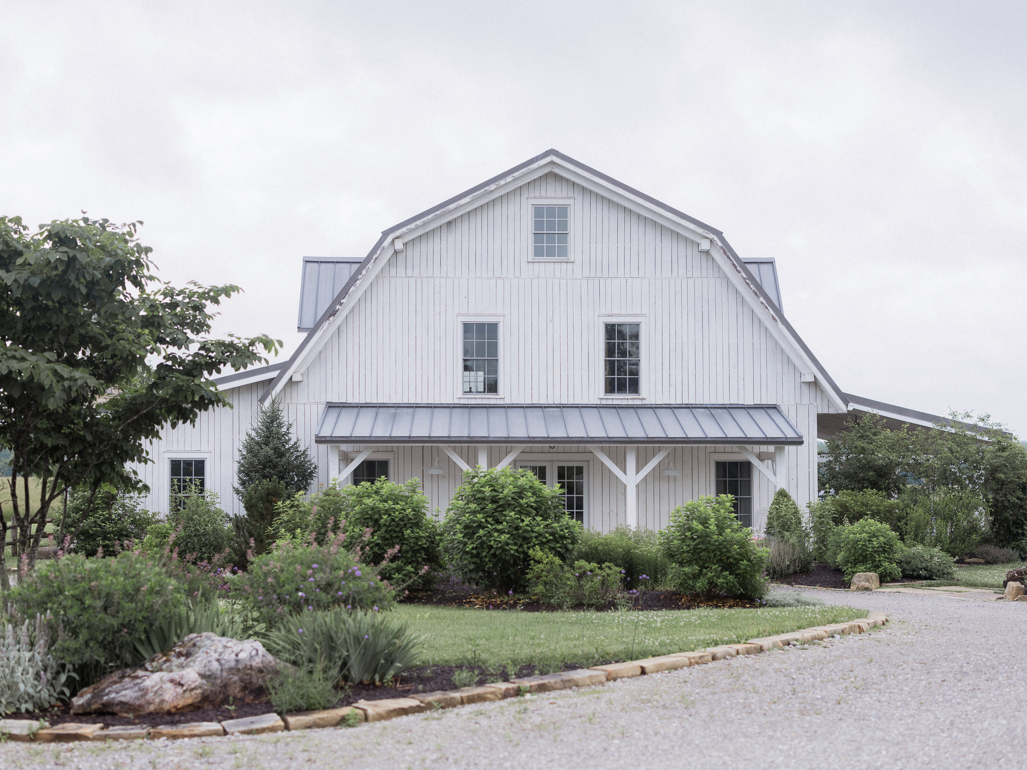 Wide shot of the white farmhouse-style barn at Blue Bell Farm in Fayette, Missouri, surrounded by lush greenery on an overcast day.