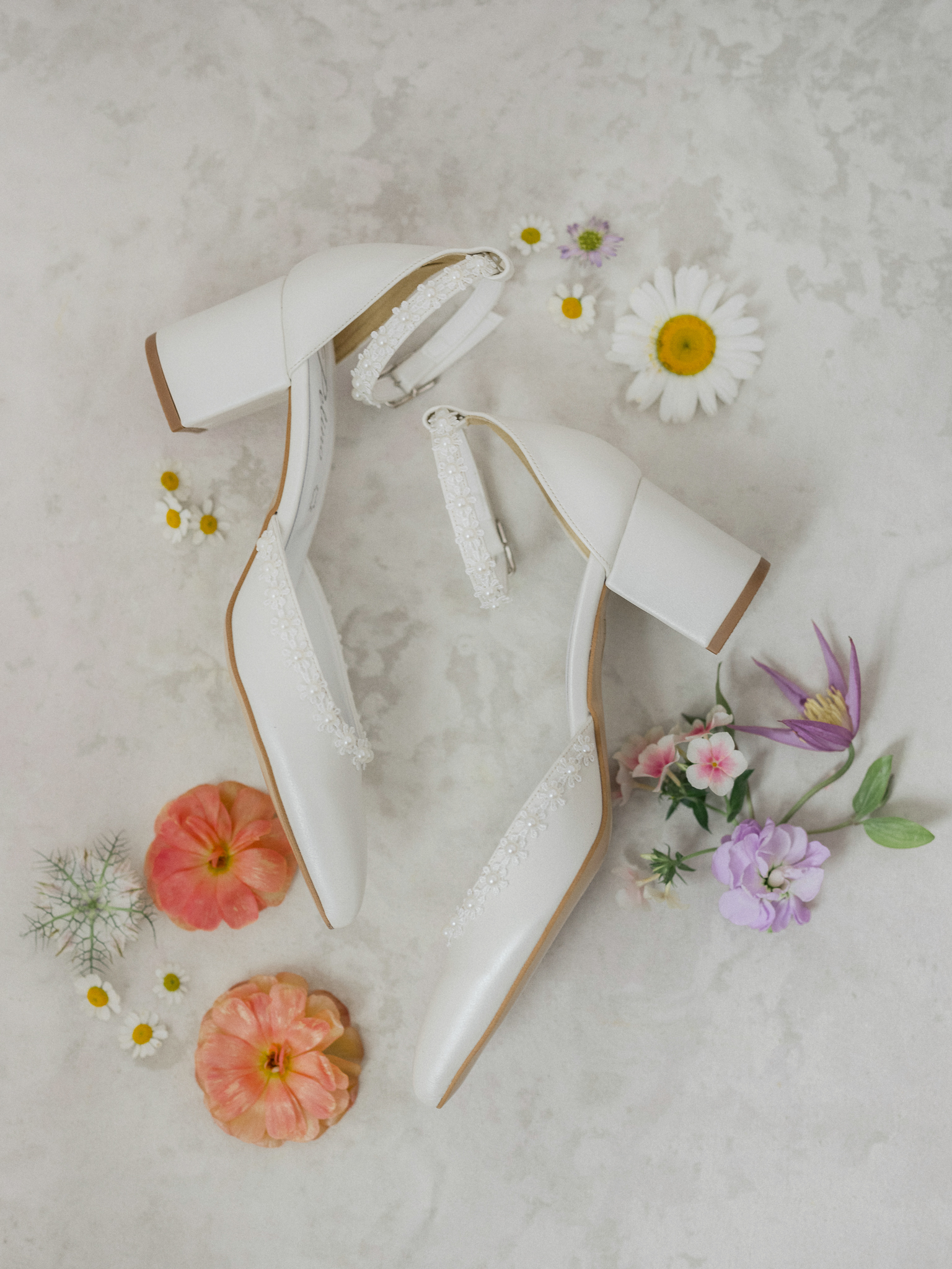 Flat lay of the bride’s white block heels with pearl ankle straps, styled with colorful wildflowers on a light background.