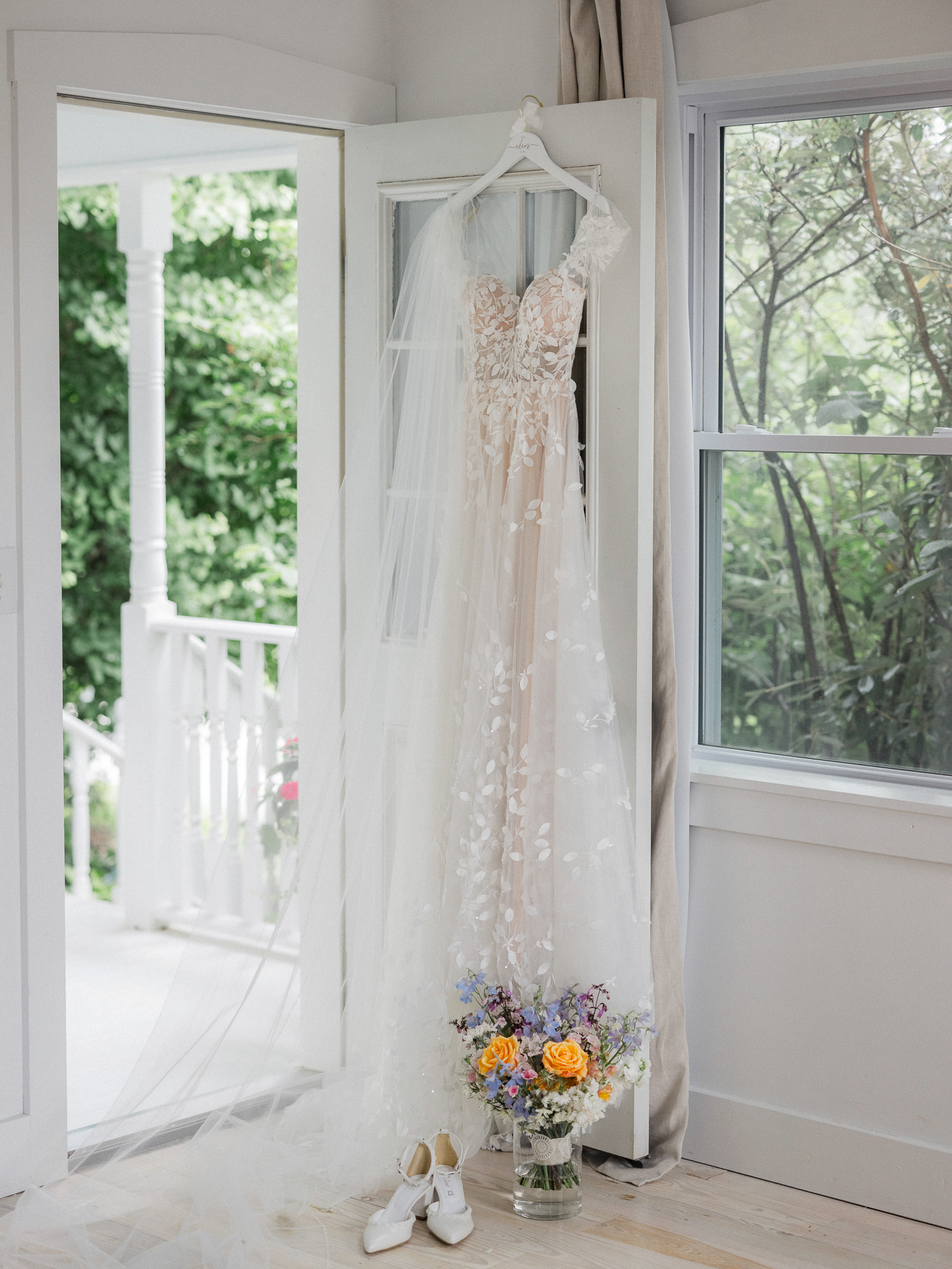 Bride’s floral lace wedding gown hanging on a door with a long veil, white heels, and bouquet placed below.
