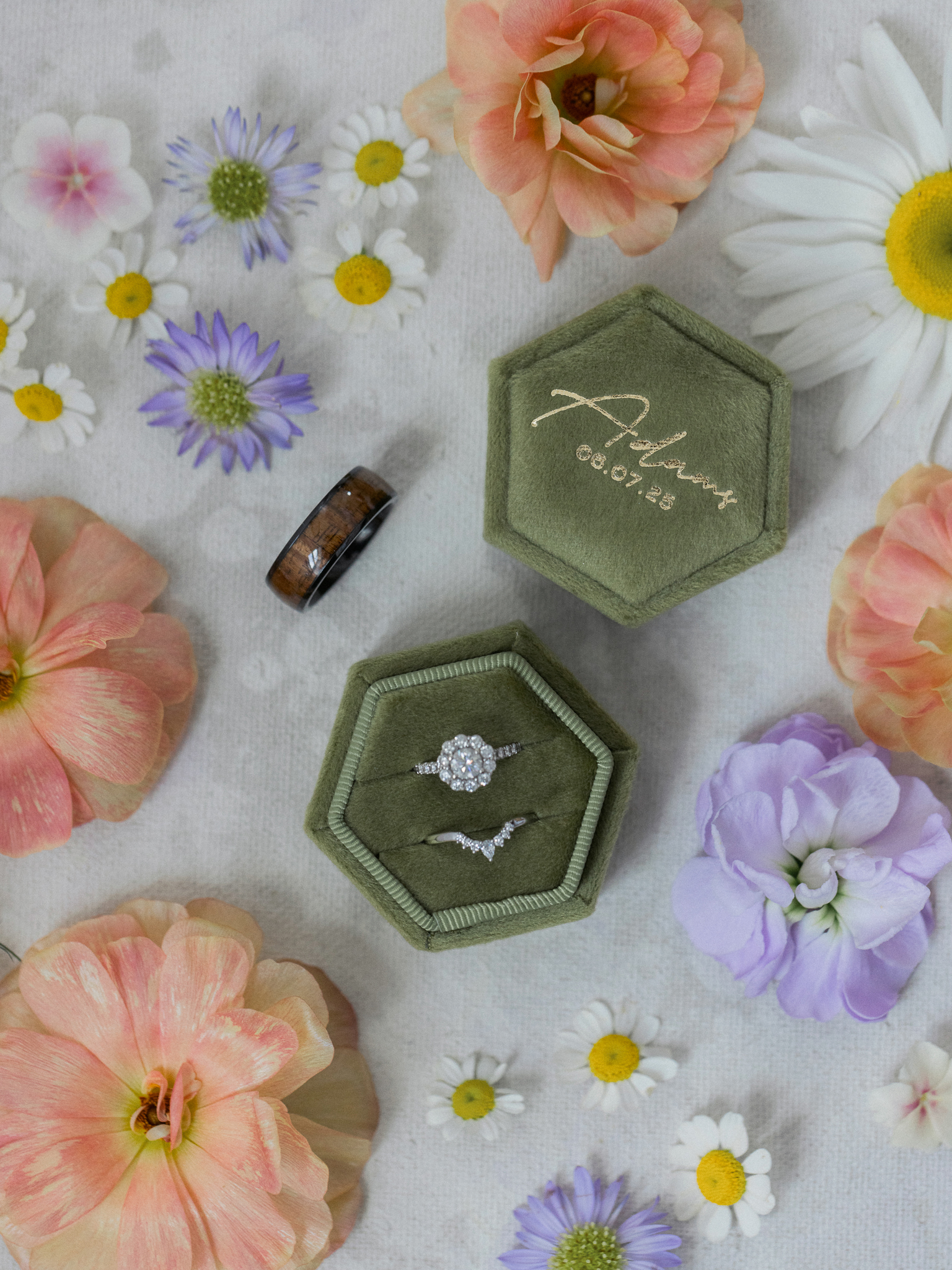 Velvet green hexagon ring box with diamond engagement and wedding rings, surrounded by pastel flowers and groom’s wood inlay band.