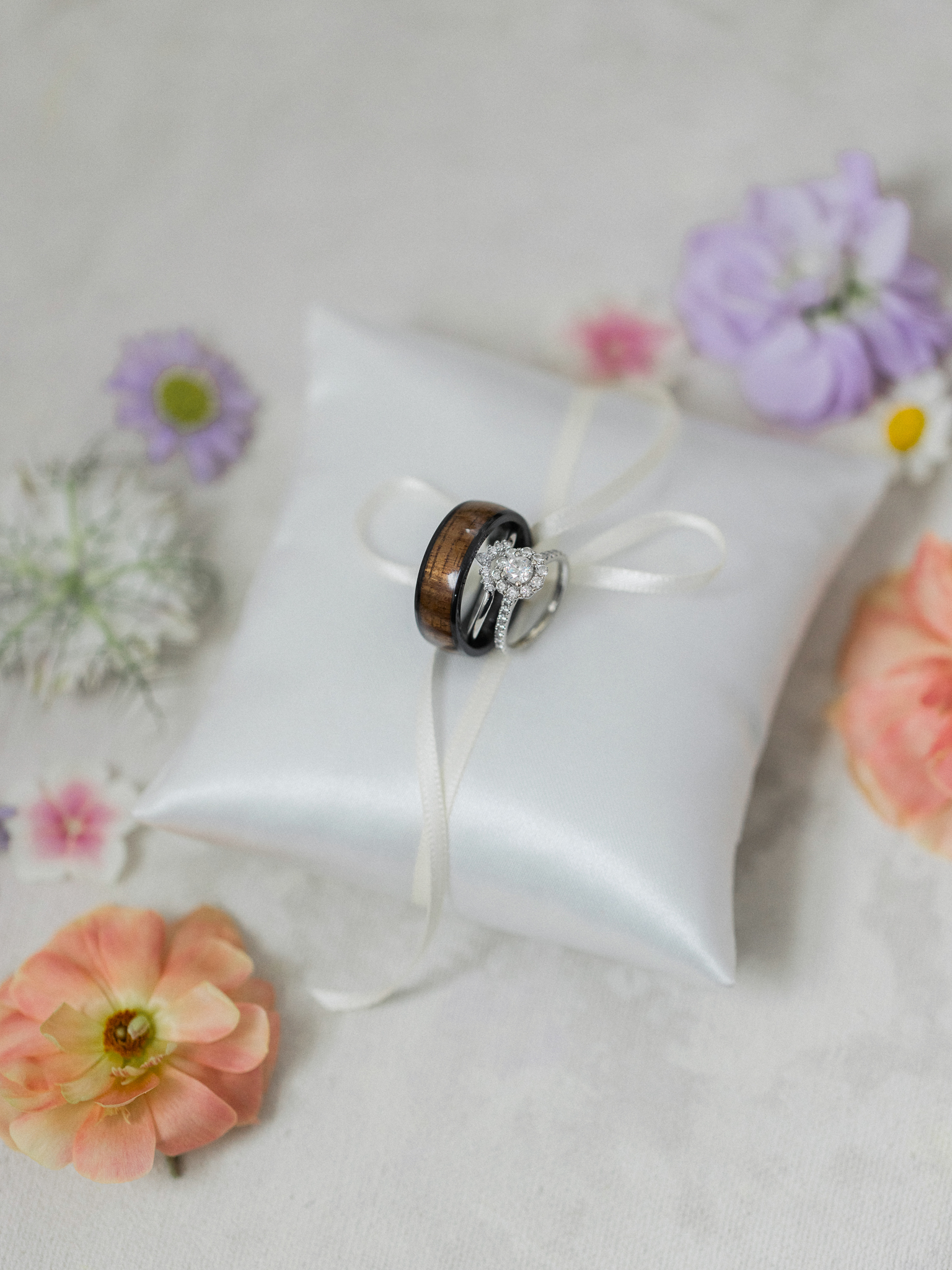 His and hers wedding rings tied with ribbon on a white satin pillow, surrounded by fresh peach, lavender, and white flowers.