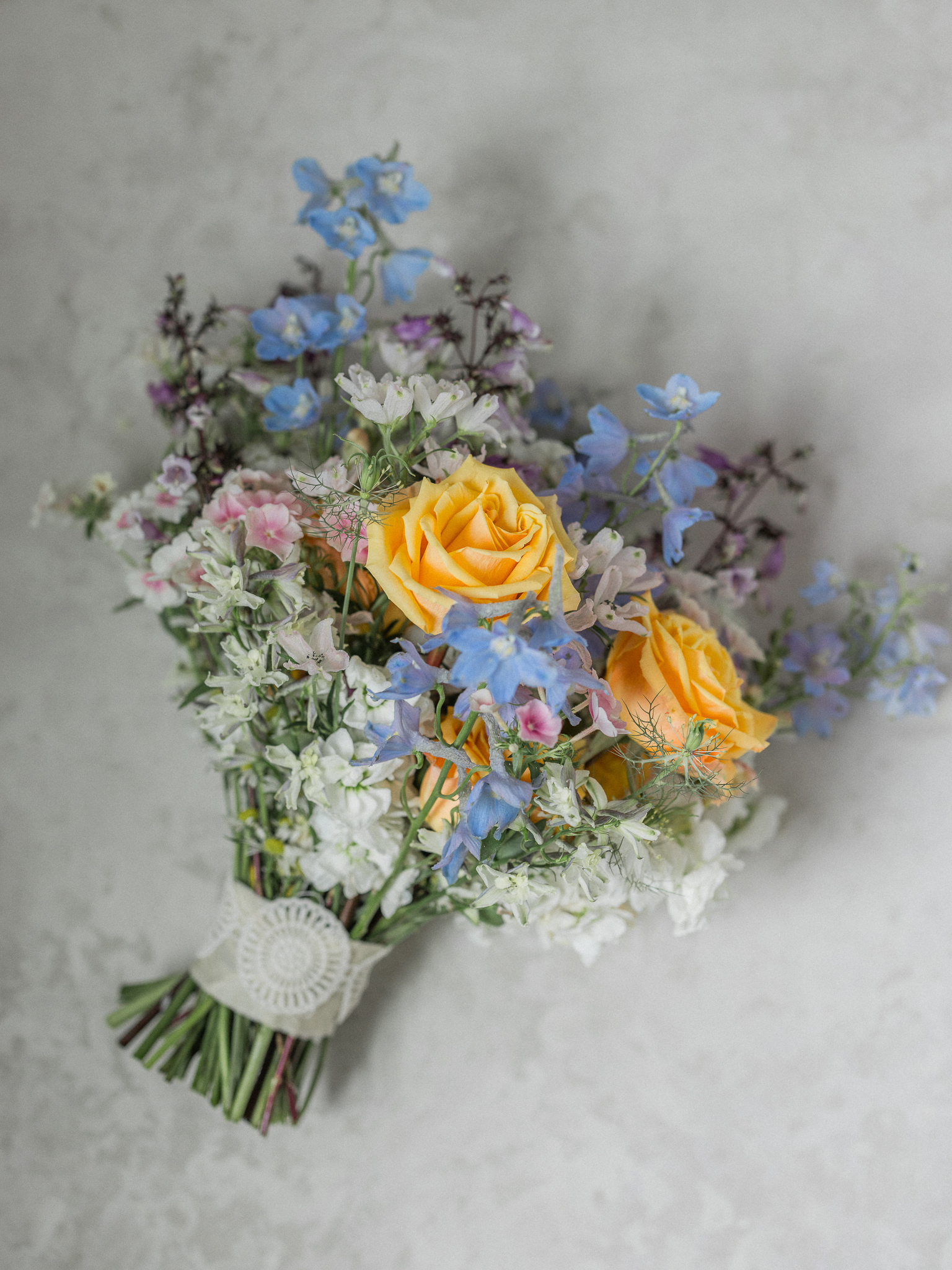 Bride’s hand-tied bouquet with yellow roses, blue delphinium, and pastel wildflowers, wrapped in a lace ribbon designed by Sugarberry Blooms.