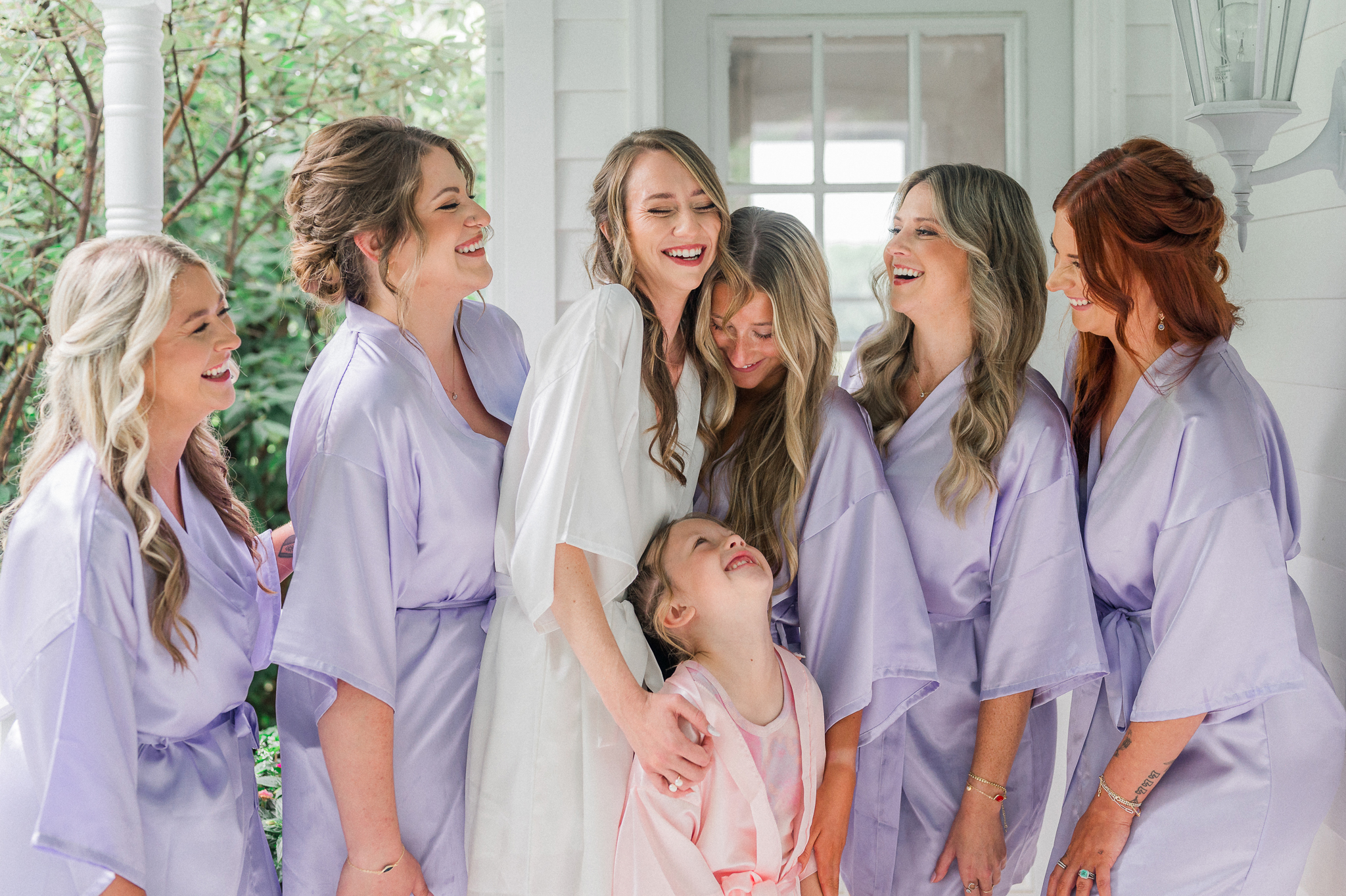 Bride laughing joyfully surrounded by bridesmaids in lavender robes and a flower girl in pink.