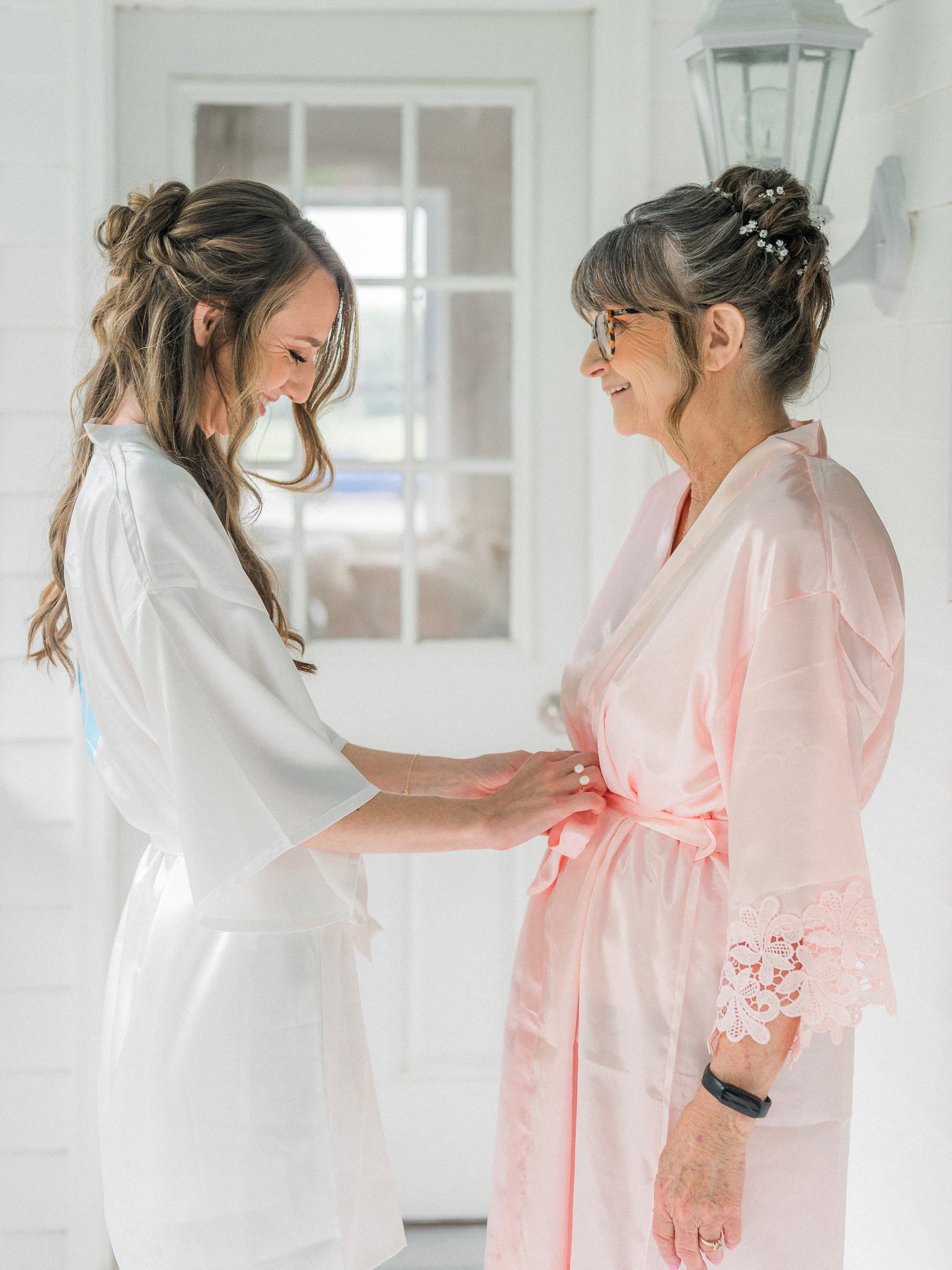 Candid moment of the bride smiling as she ties the sash on her mother’s blush pink robe during wedding morning prep.