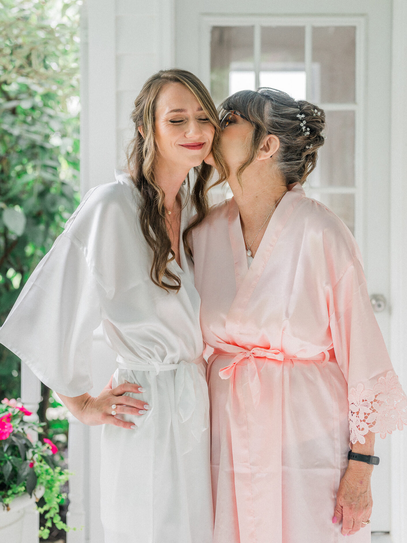 Bride in a white robe receives a kiss on the cheek from her mother in matching blush robe, standing together on a porch.