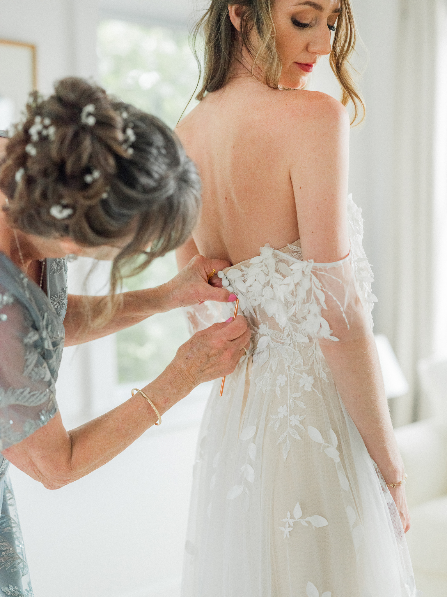 Mother of the bride carefully fastening the back of the bride’s dress in a bright getting-ready room.