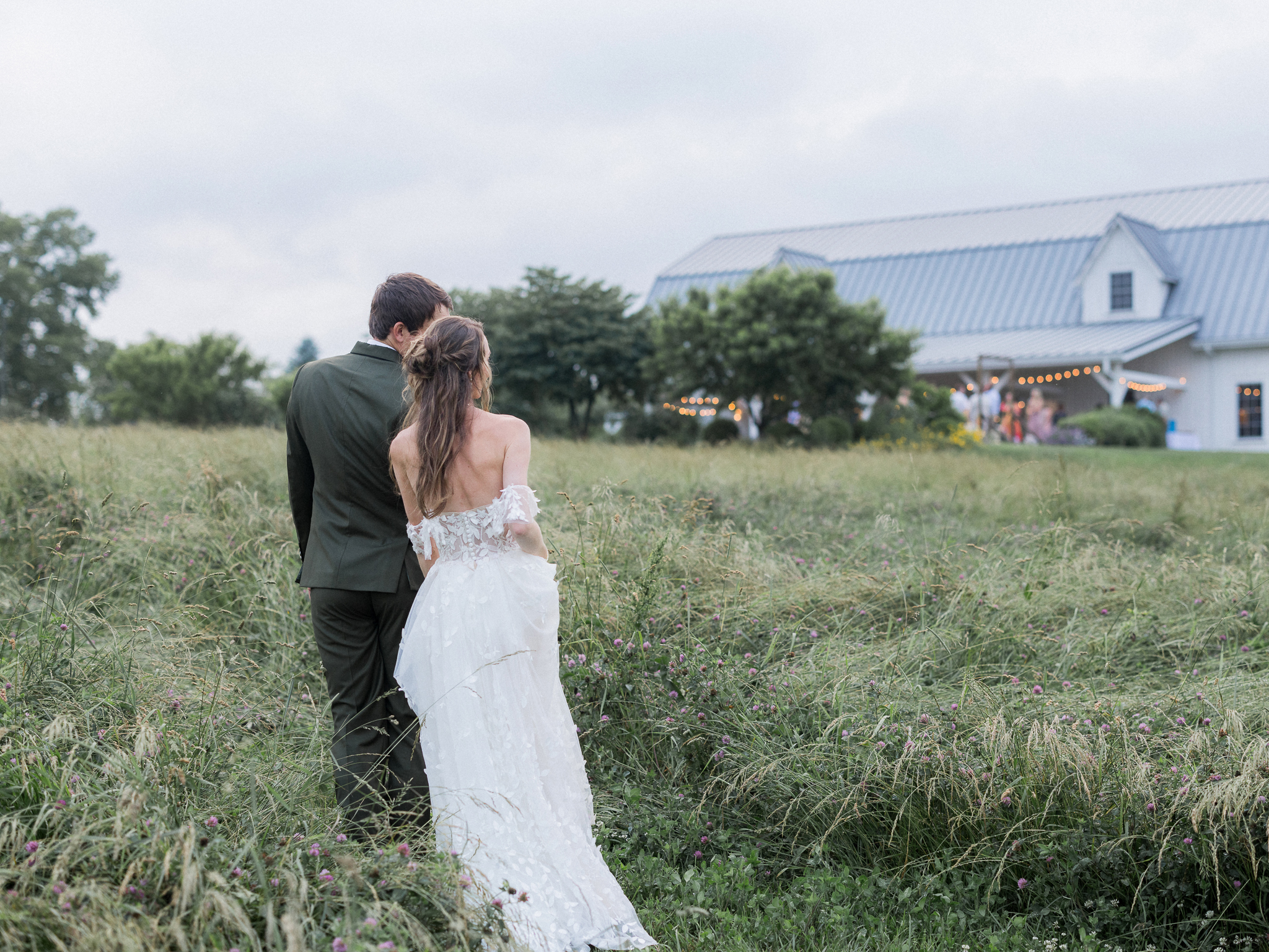 Bride and groom walk through a grassy field at Blue Bell Farm toward their warmly lit reception barn in the distance, captured from behind.