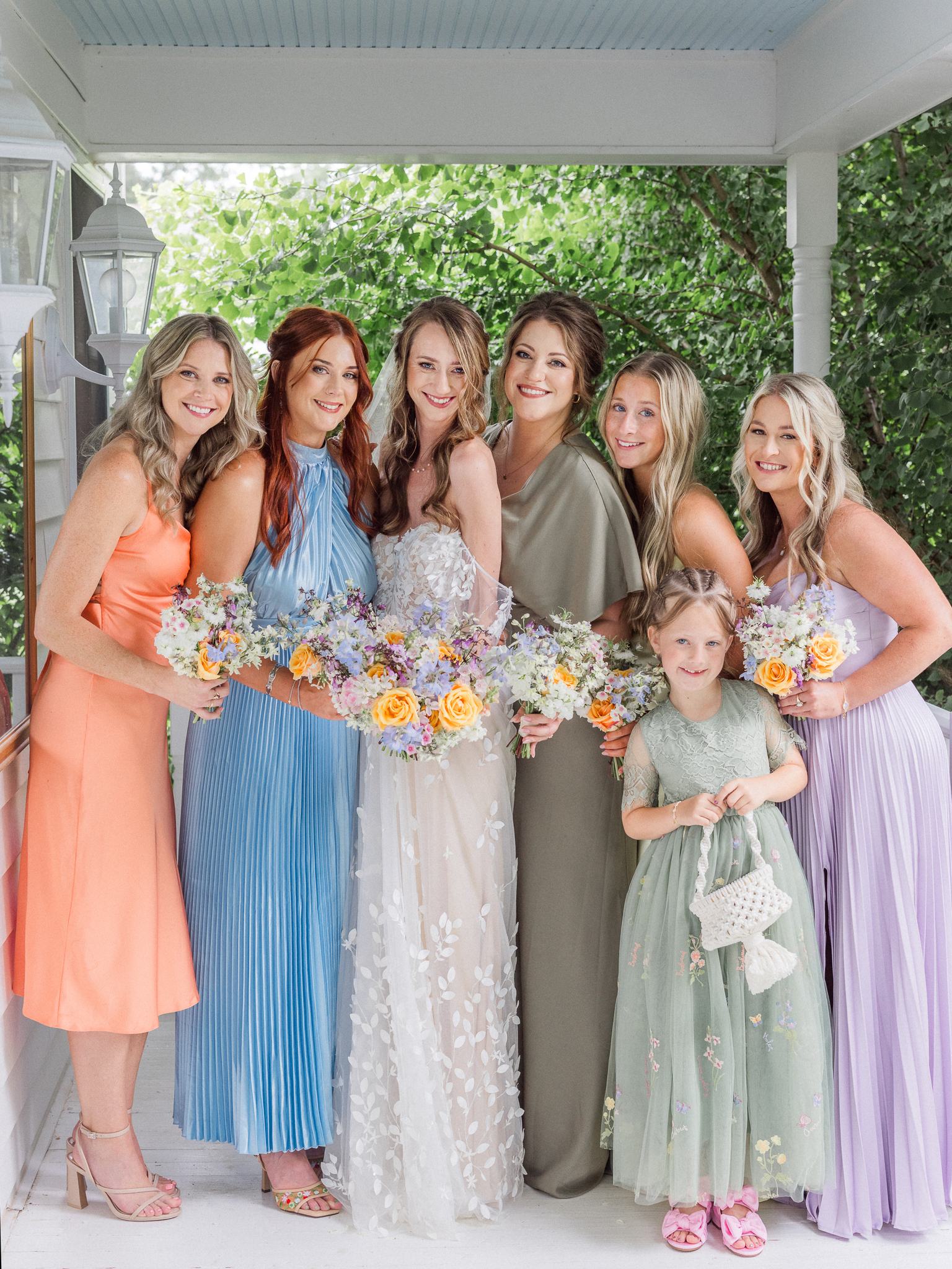 Bride posing with bridesmaids and flower girl in colorful dresses and holding bright floral bouquets on porch of Blue Bell Farm.