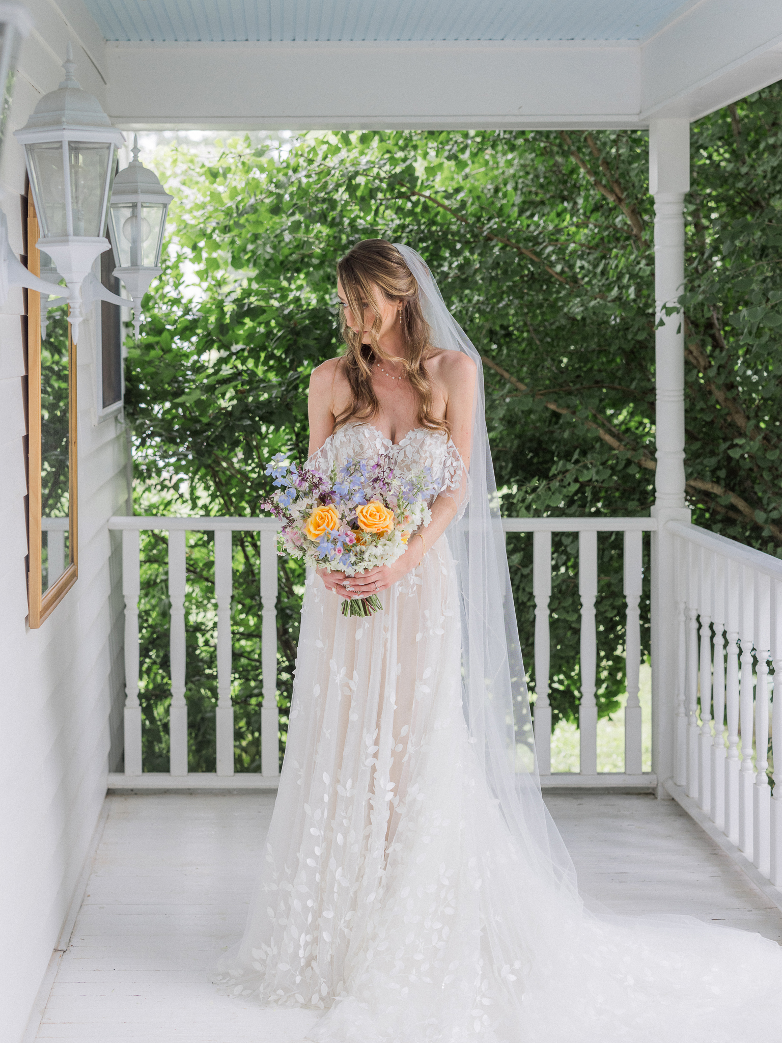 Bride standing alone on white farmhouse porch with wildflower bouquet and flowing floral lace gown.