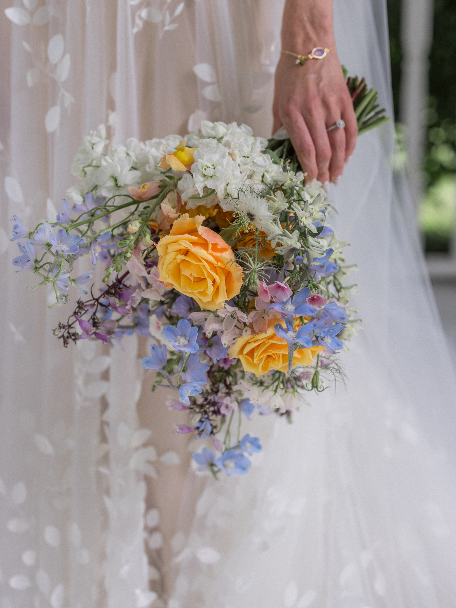Close-up of bride’s hand holding colorful bouquet of yellow roses, blue and white wildflowers with engagement ring visible.
