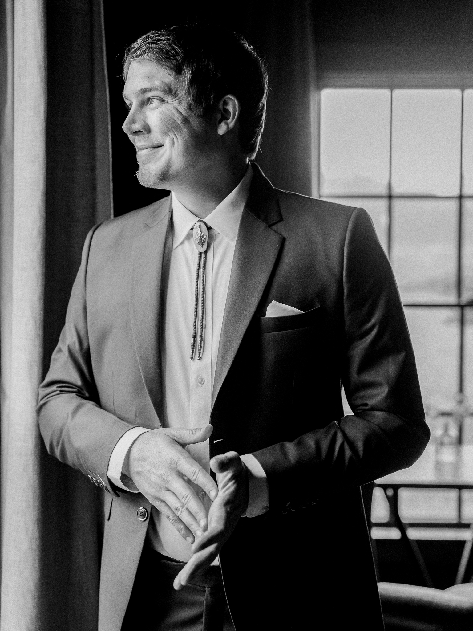 Groom smiling and adjusting his sleeves near a window, wearing a green suit and bolo tie, shot by Love Tree Studios.