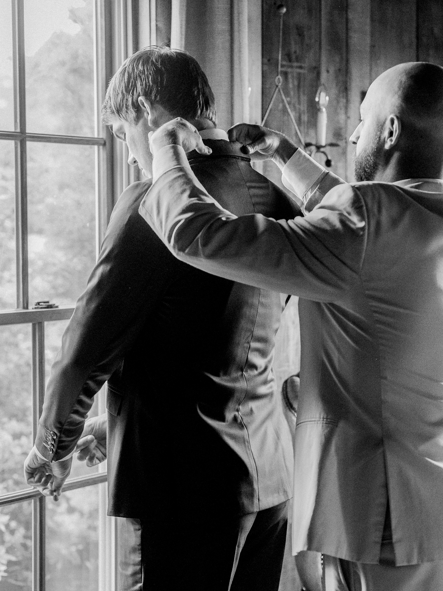 Groomsman helping groom adjust his collar near large rustic windows with natural light streaming in.
