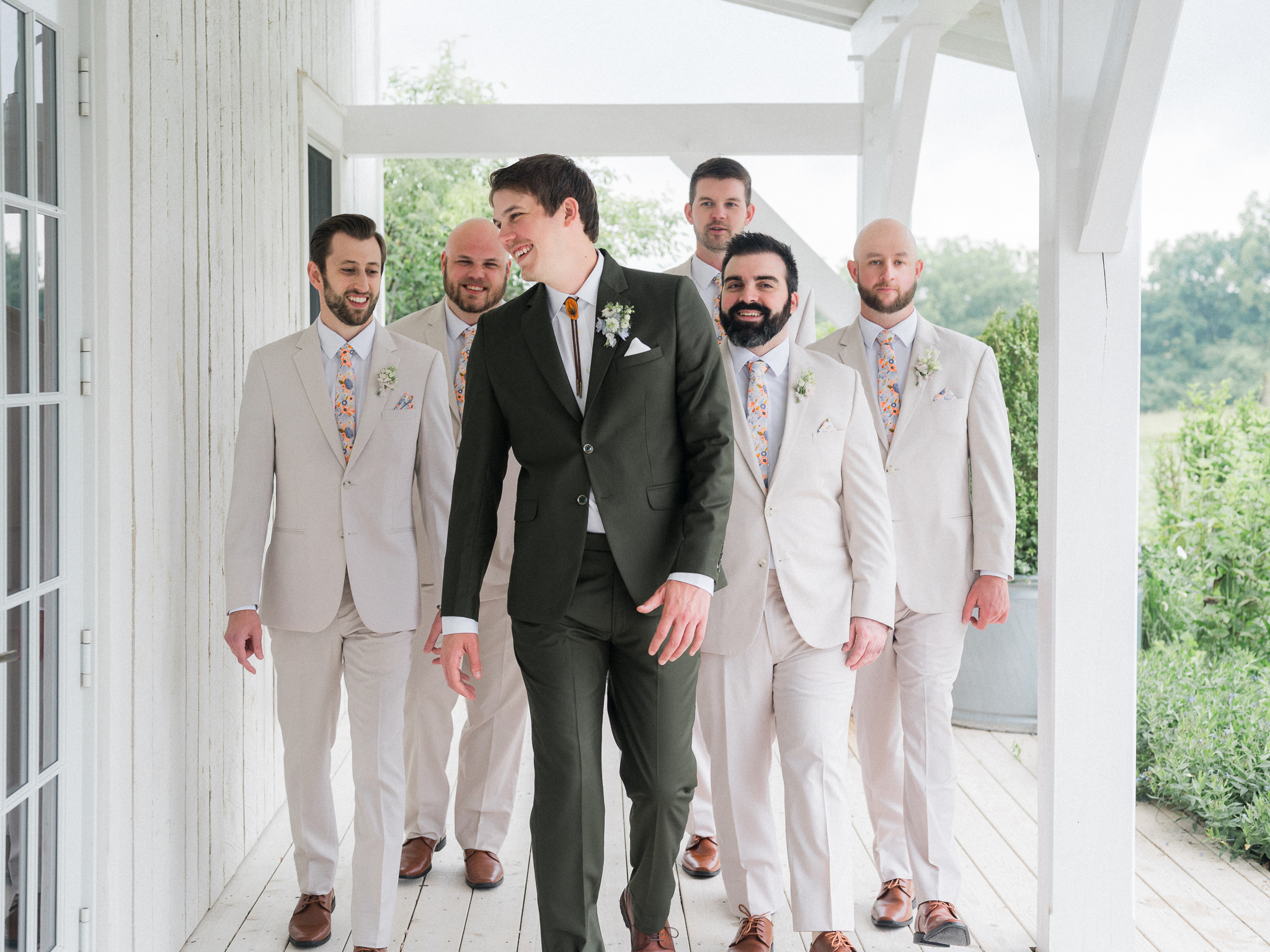 Groom walking confidently in front of groomsmen in cream-colored suits on porch at Blue Bell Farm.