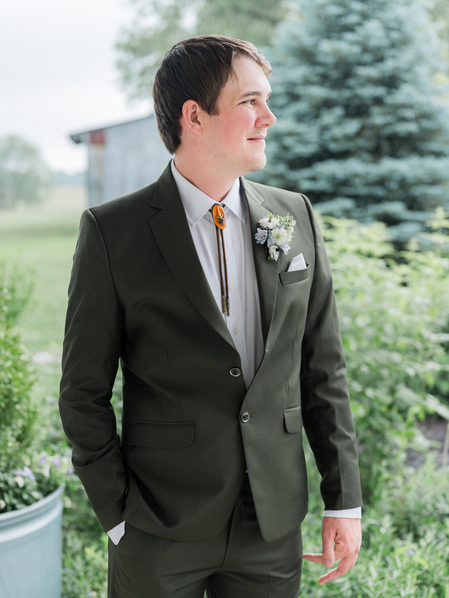 Groom smiling in portrait, wearing a dark green suit and boutonniere outside on a covered porch.
