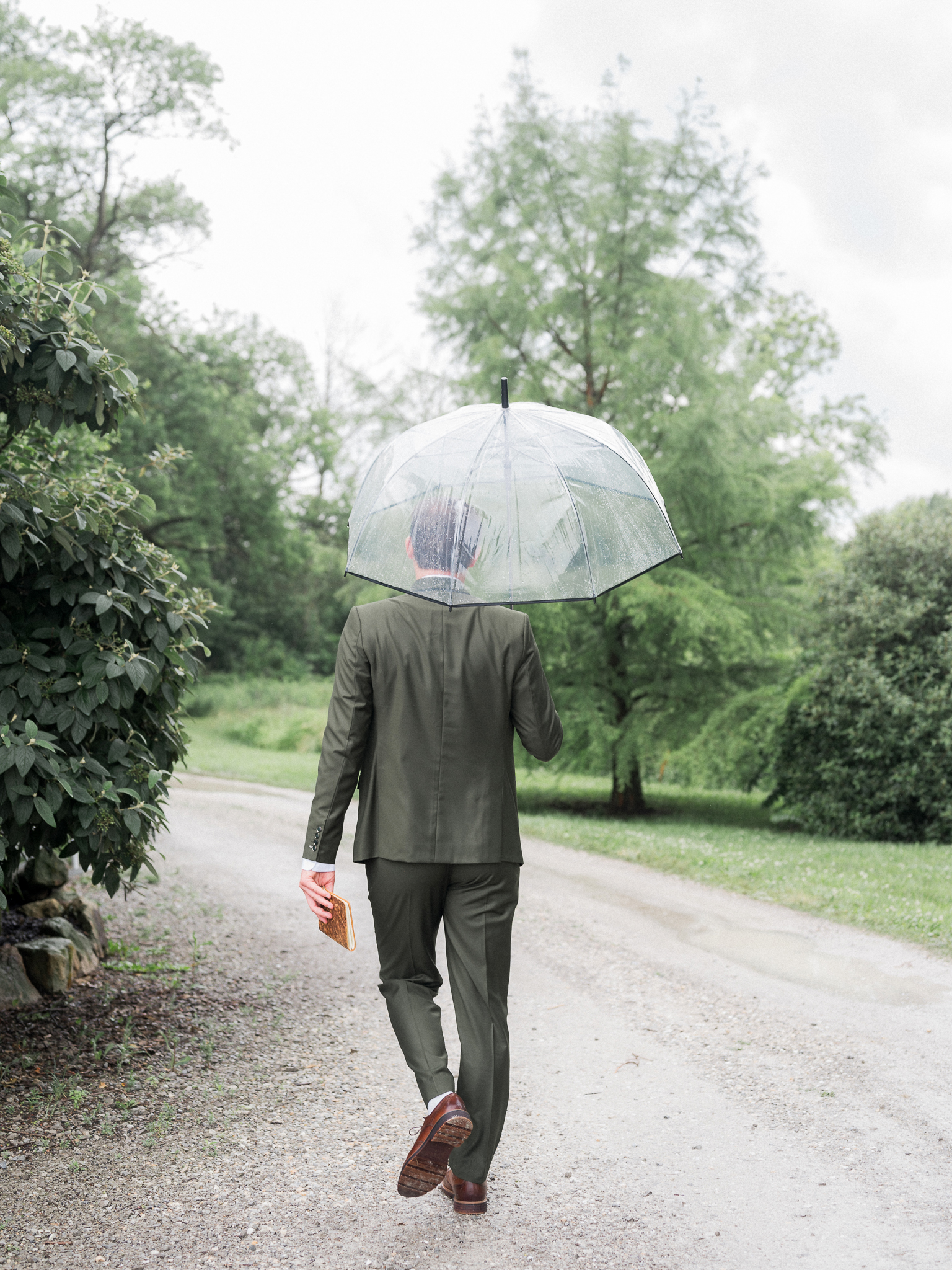 Groom walking down gravel path with clear umbrella in hand, heading toward ceremony location at Blue Bell Farm.