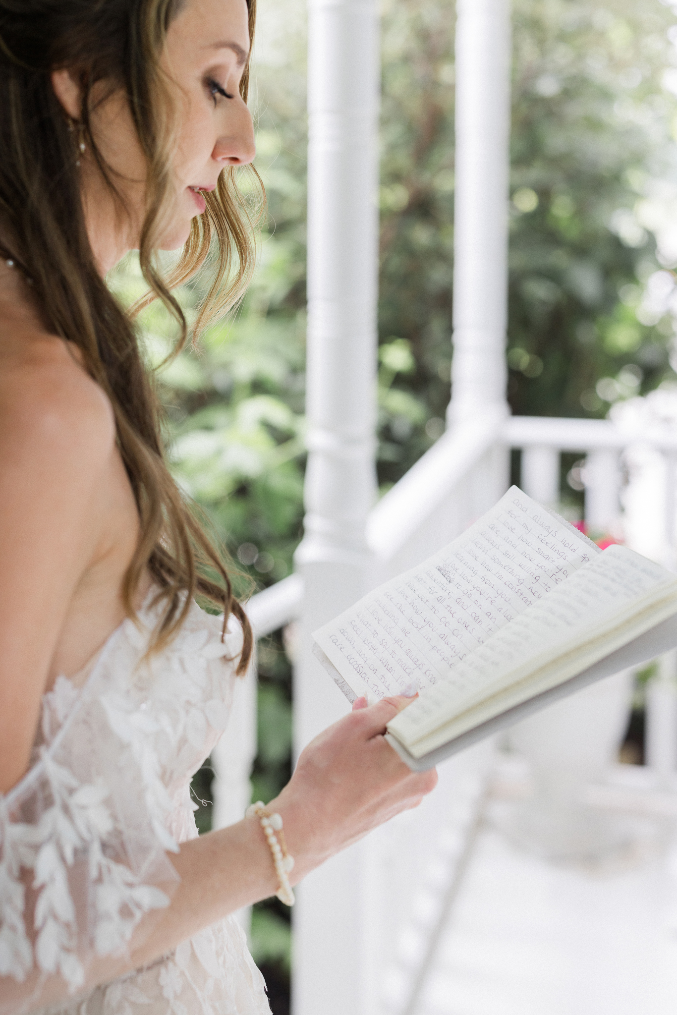 Bride reading handwritten vows from notebook while standing on white porch surrounded by greenery at Blue Bell Farm.