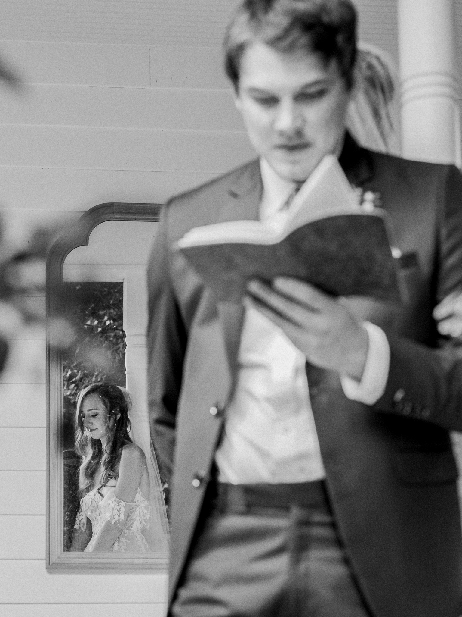 Groom reads a vow book while bride’s reflection appears in a mirror behind him at a Blue Bell Farm wedding.