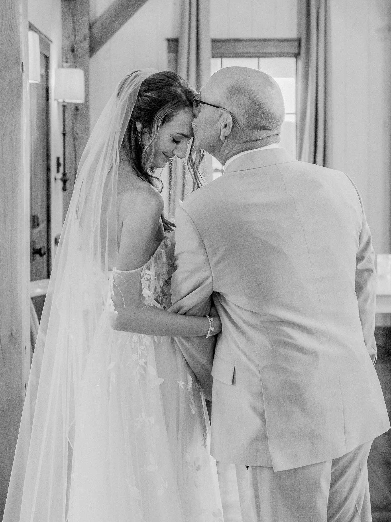 Bride smiles as her father kisses her head in a tender moment indoors at a Blue Bell Farm wedding.