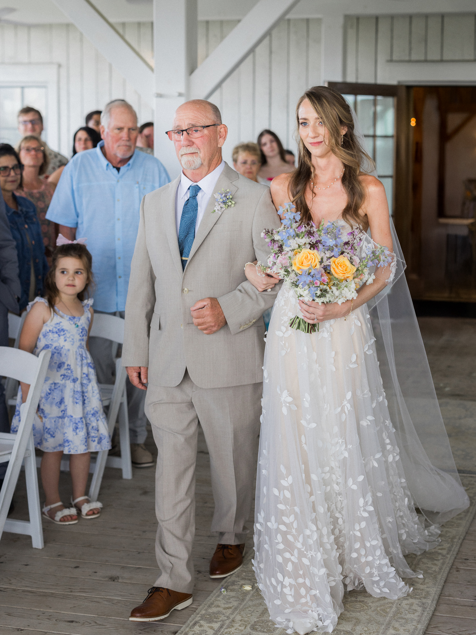 Bride walks arm-in-arm with her father, holding her bouquet while guests look on during a Blue Bell Farm wedding.