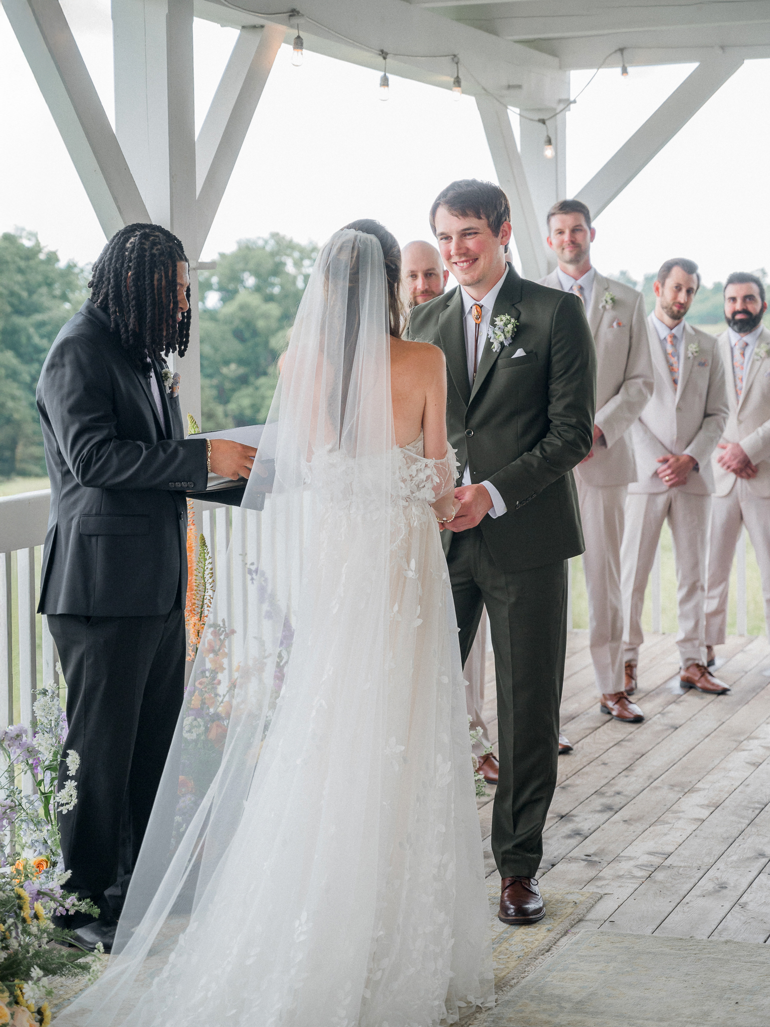 Groom holds bride’s hands and smiles during the wedding ceremony with groomsmen in view at a Blue Bell Farm wedding.