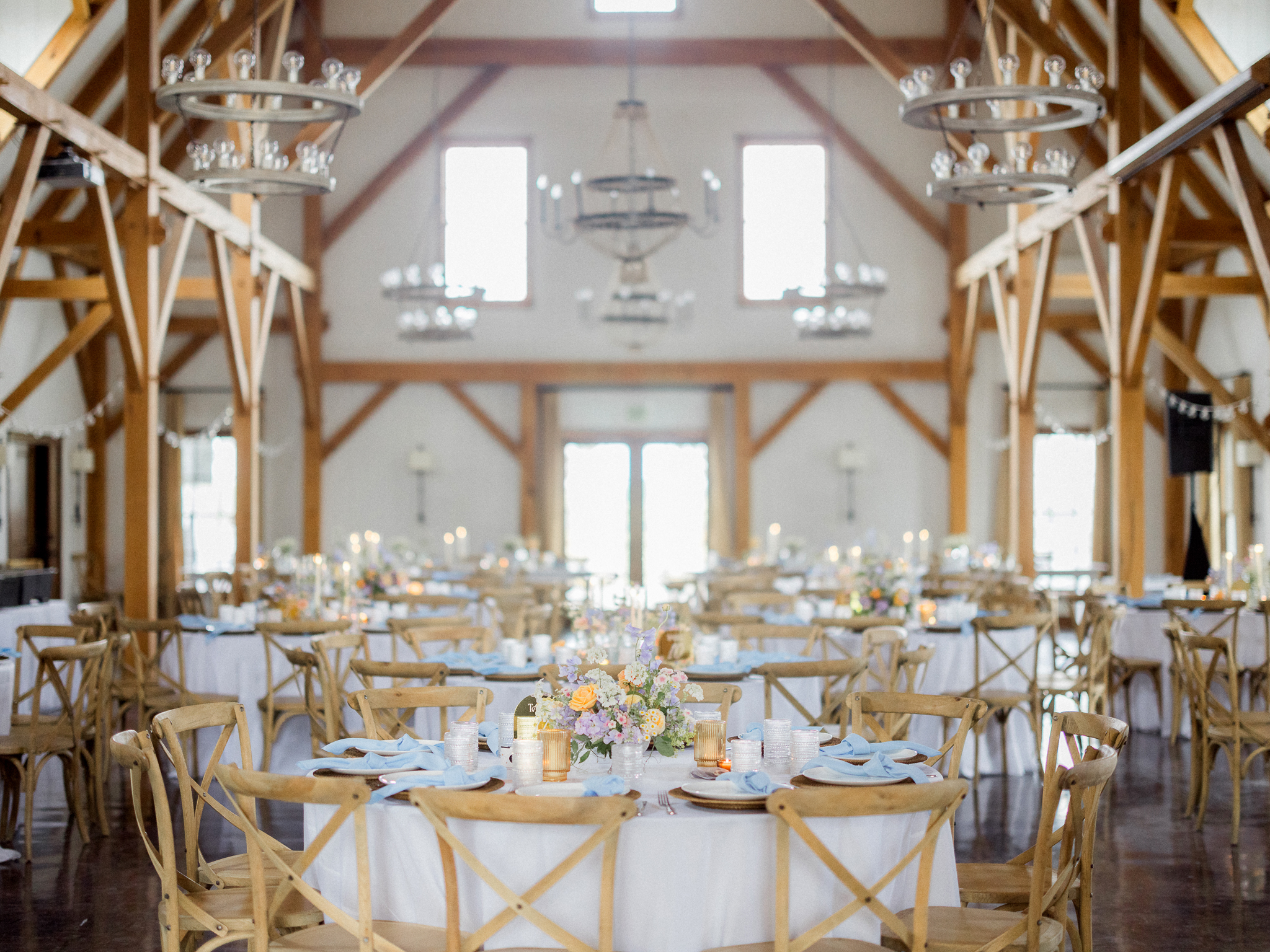 Wide shot of the reception barn set with round tables, cross-back chairs, and glowing candles.