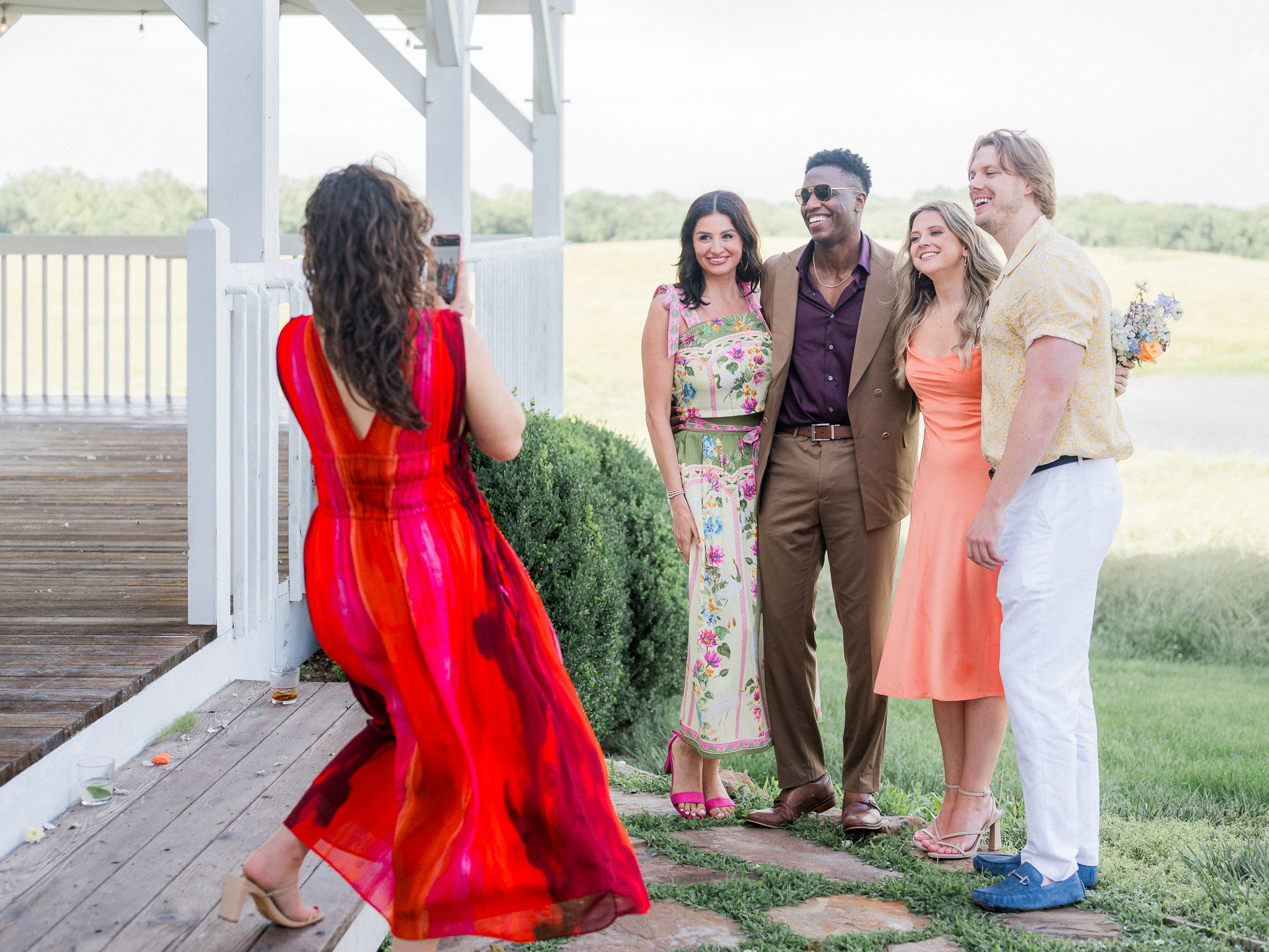 A group of four stylishly dressed wedding guests posing for a phone photo on the lawn outside the venue.